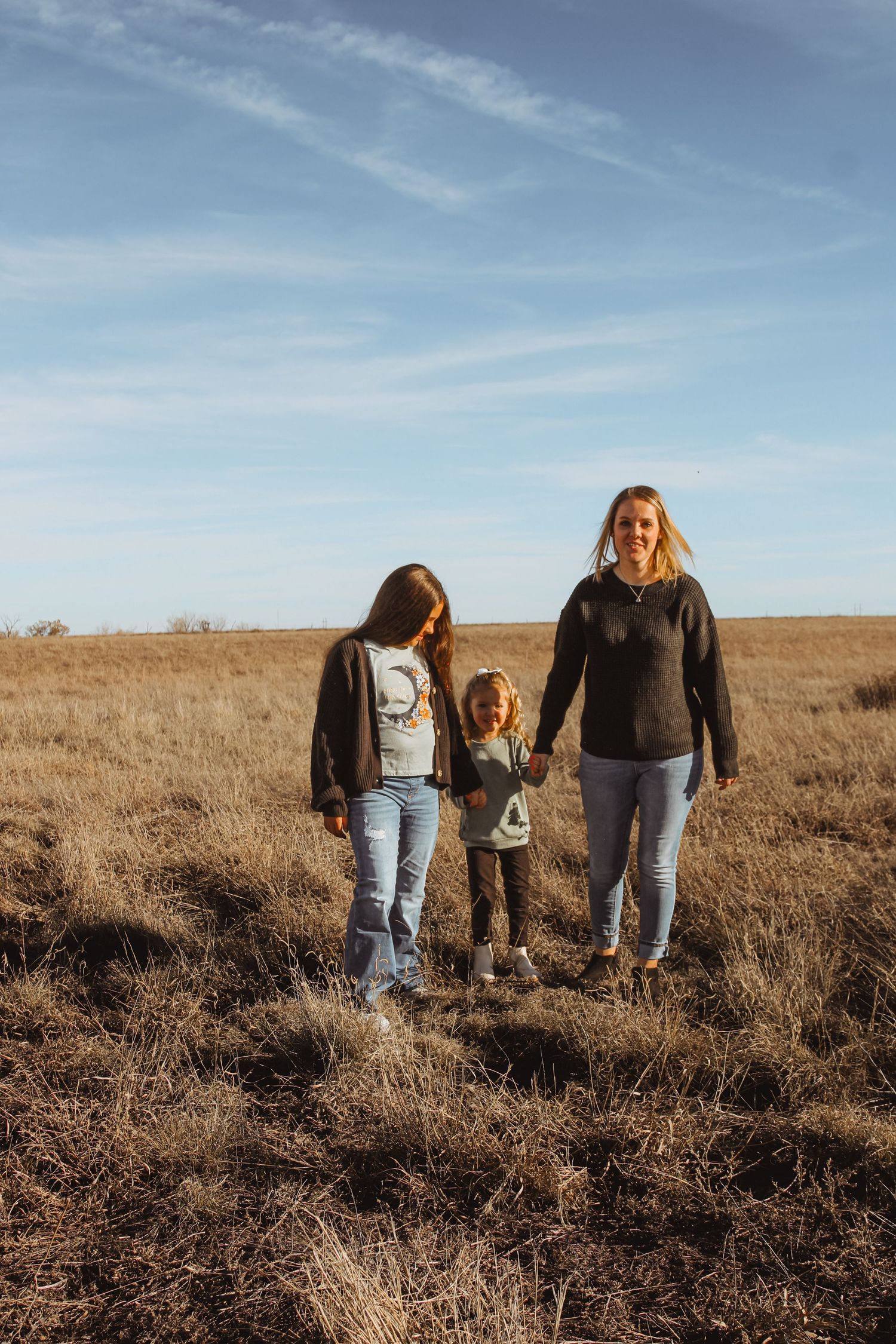 Three people walking together through a sunlit field of dry grass.