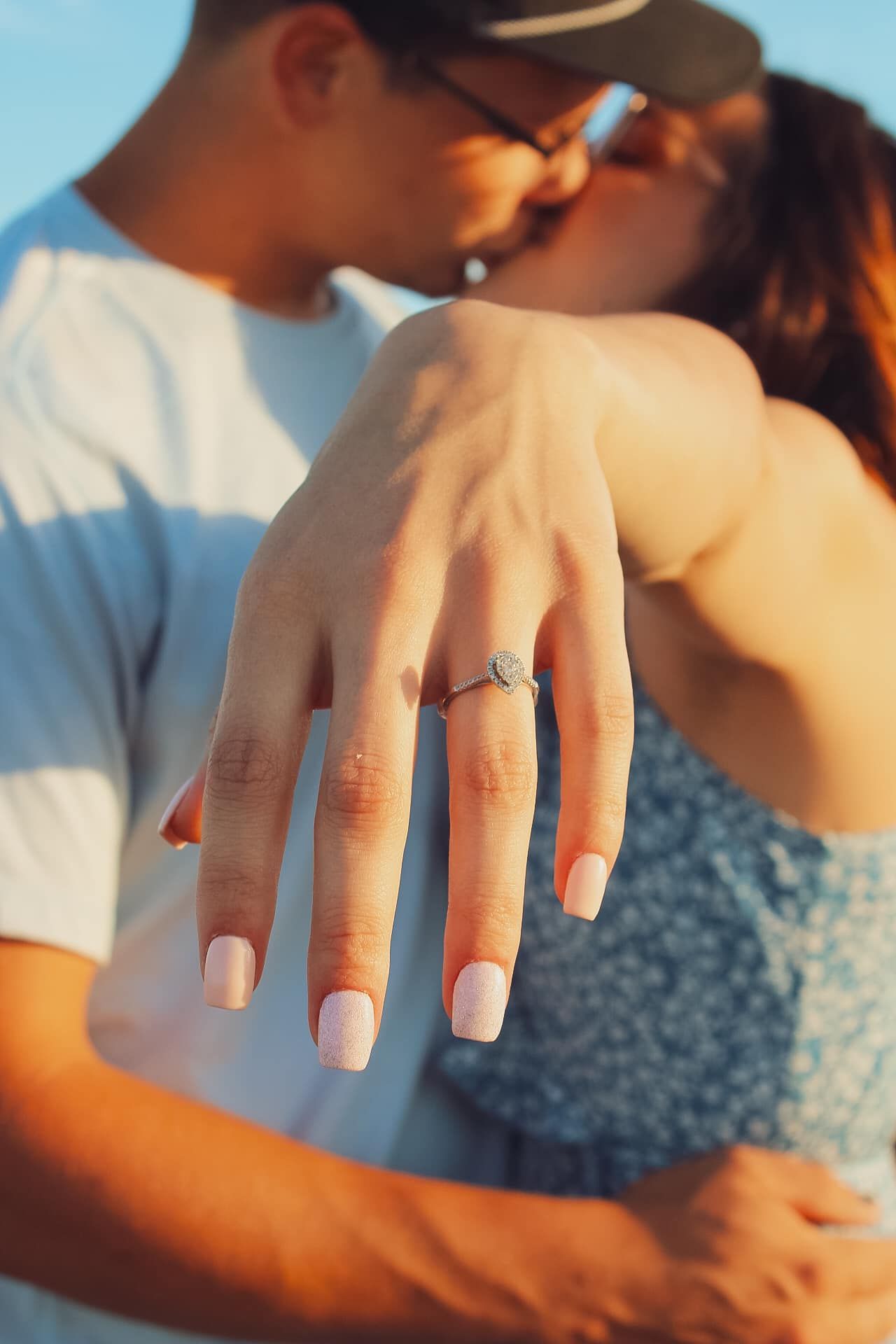 Close-up of an engagement ring on a hand with a romantic kiss in the background.