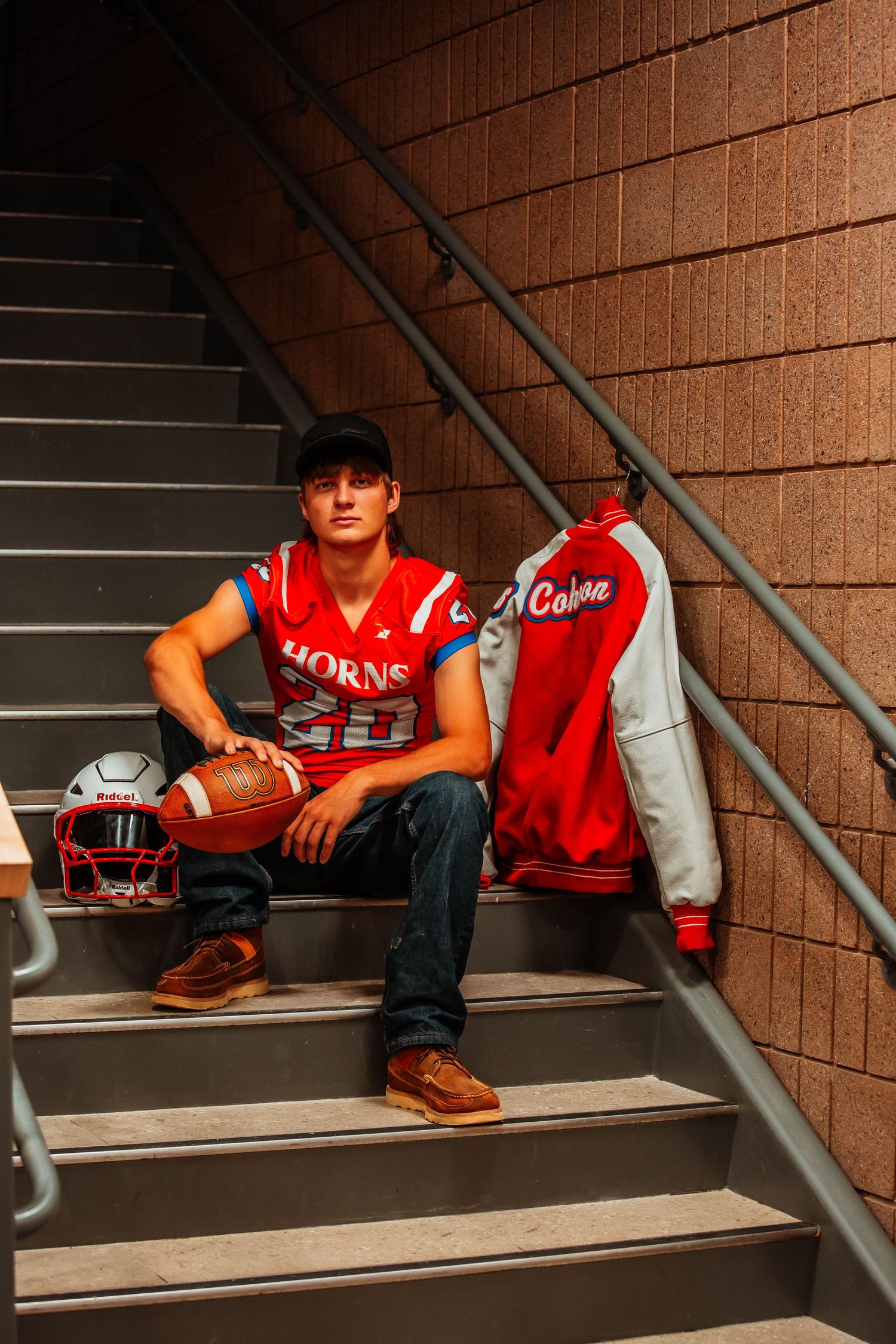 High school football player in red varsity jacket sits thoughtfully on concrete stairs with football and helmet nearby.