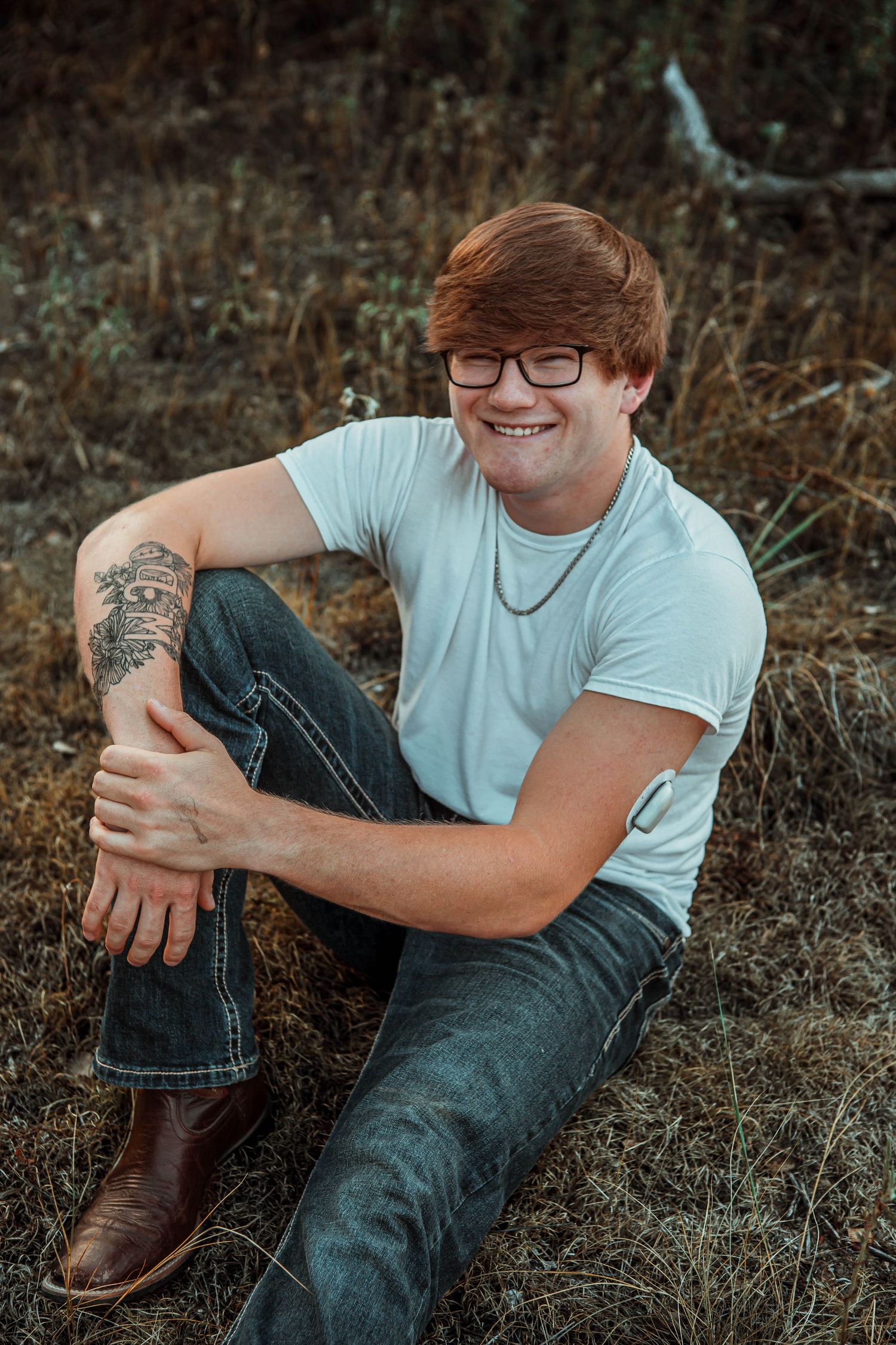 A person wearing glasses and a white t-shirt sits casually outdoors while smiling and resting their arm on their knee.