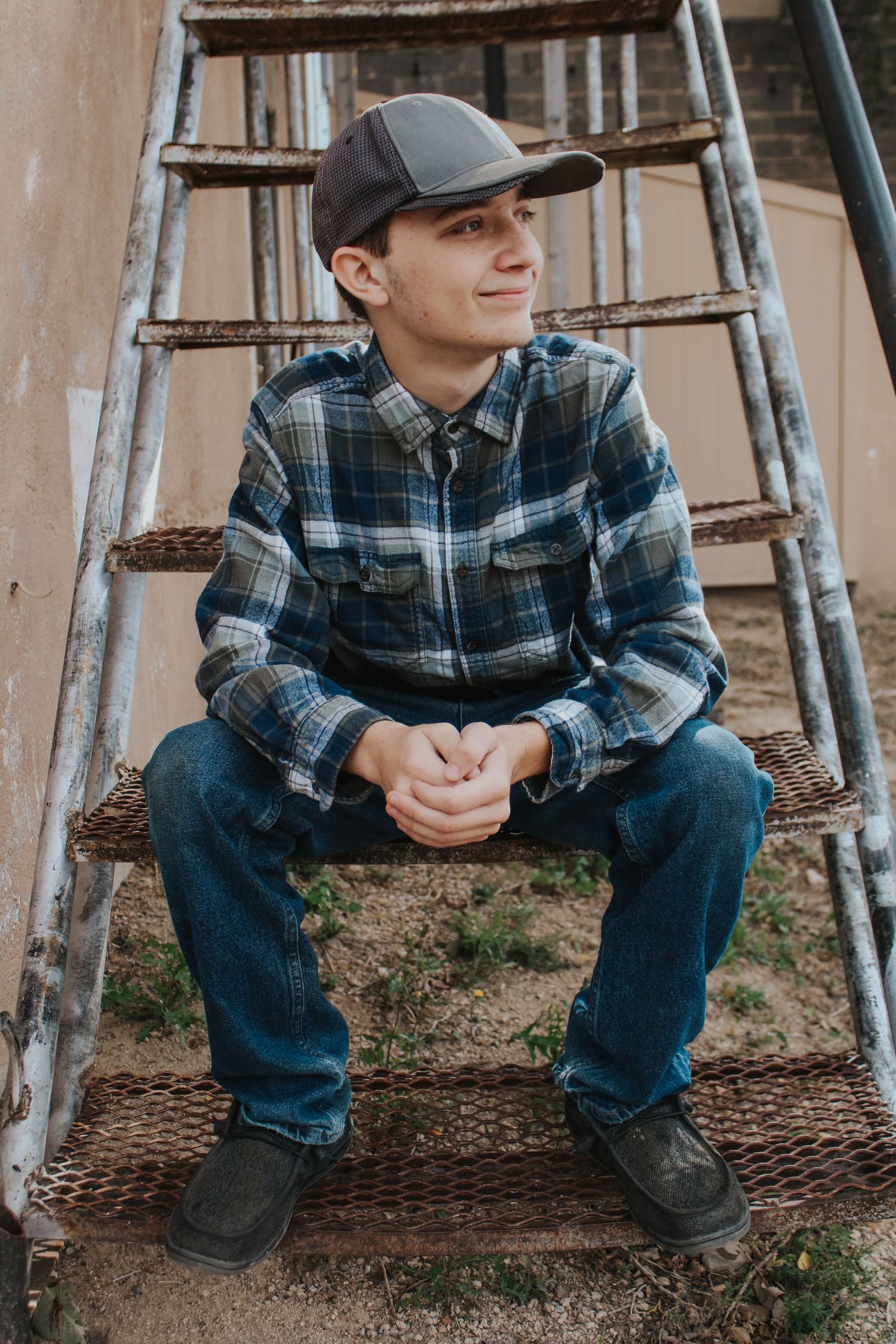 A young person in a plaid flannel shirt and baseball cap sits on rustic wooden stairs outdoors.