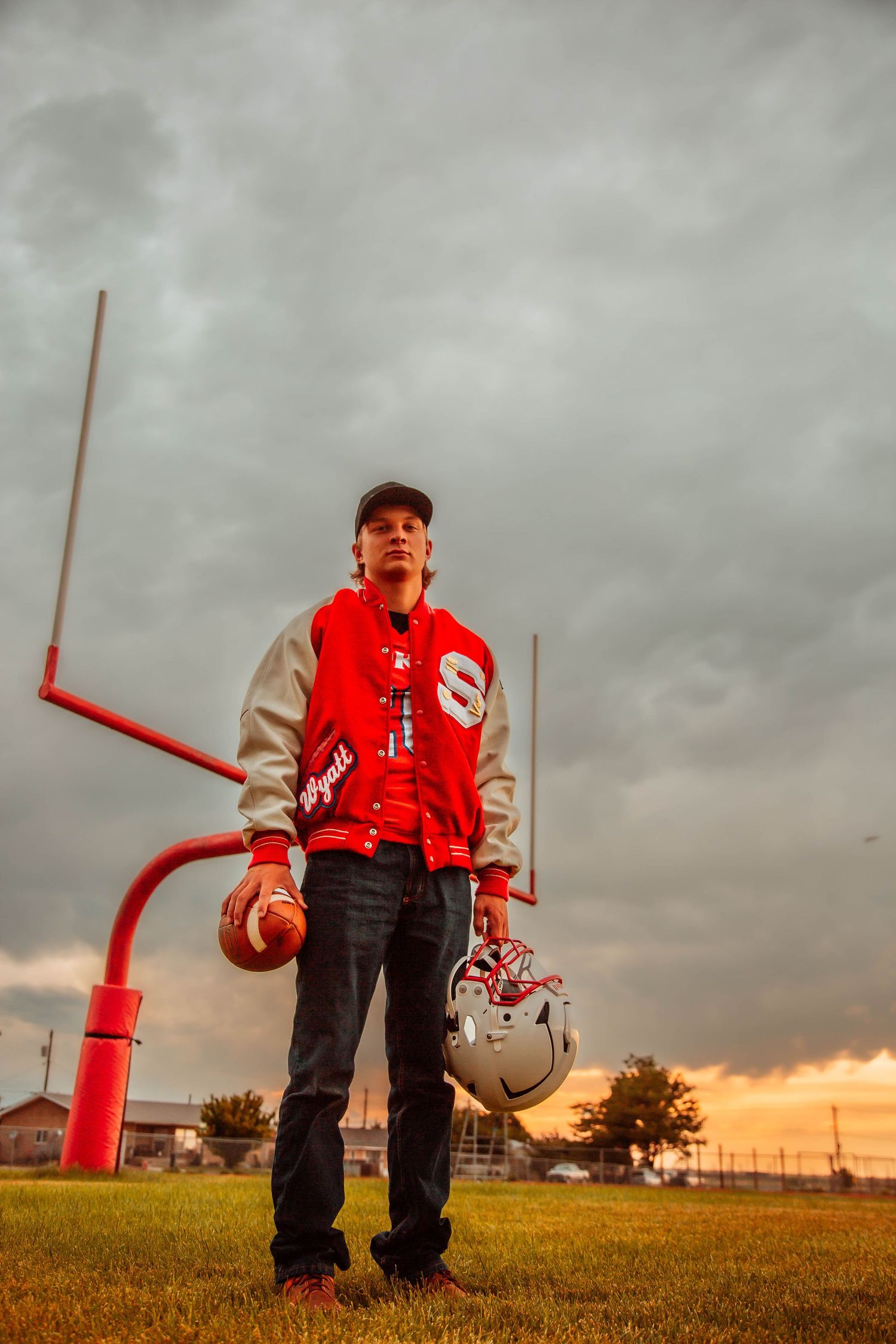 Football player stands near goal post on grassy field at sunset with dramatic cloudy sky.