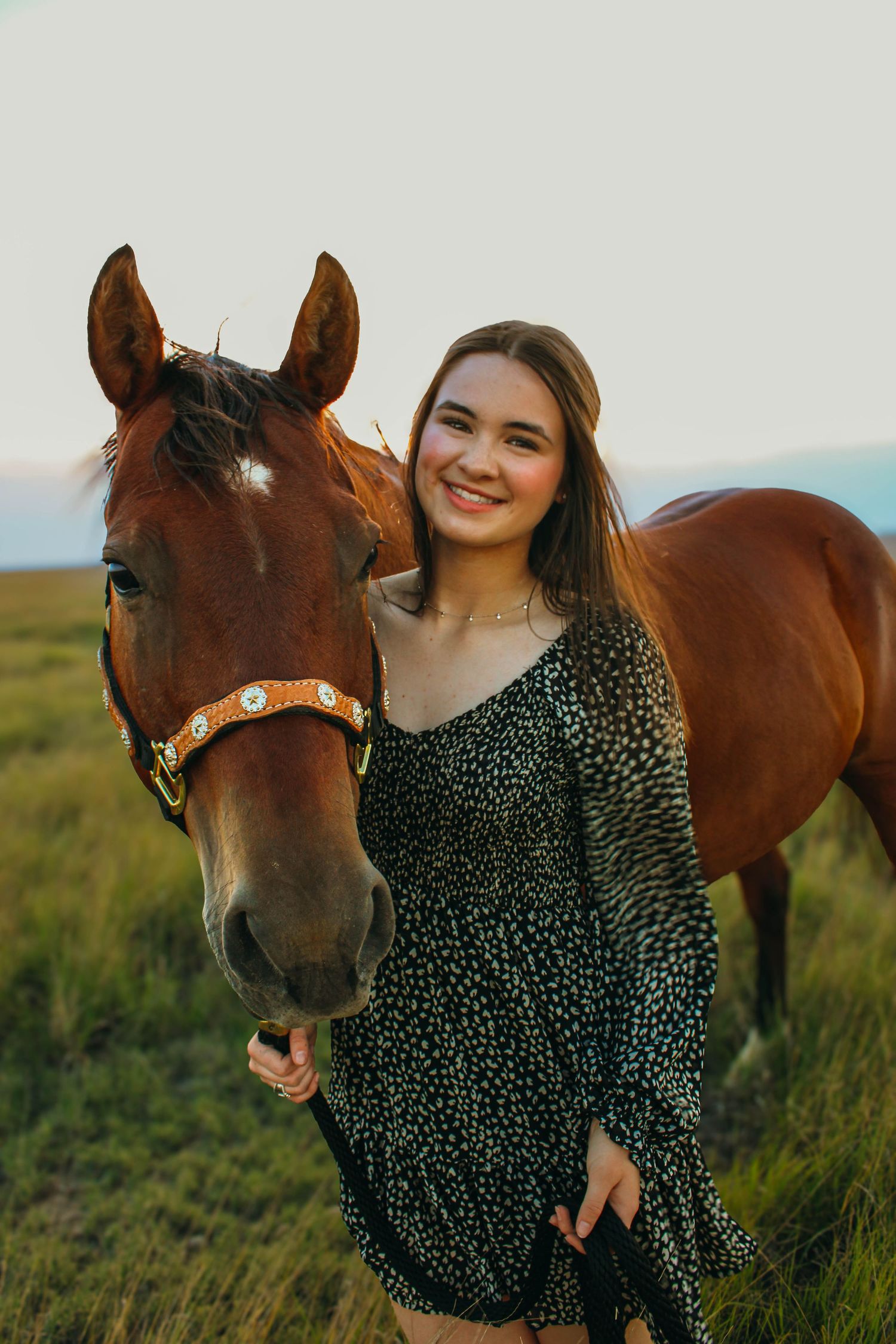 A person in a polka dot dress poses with a brown horse in a grassy field during golden hour.