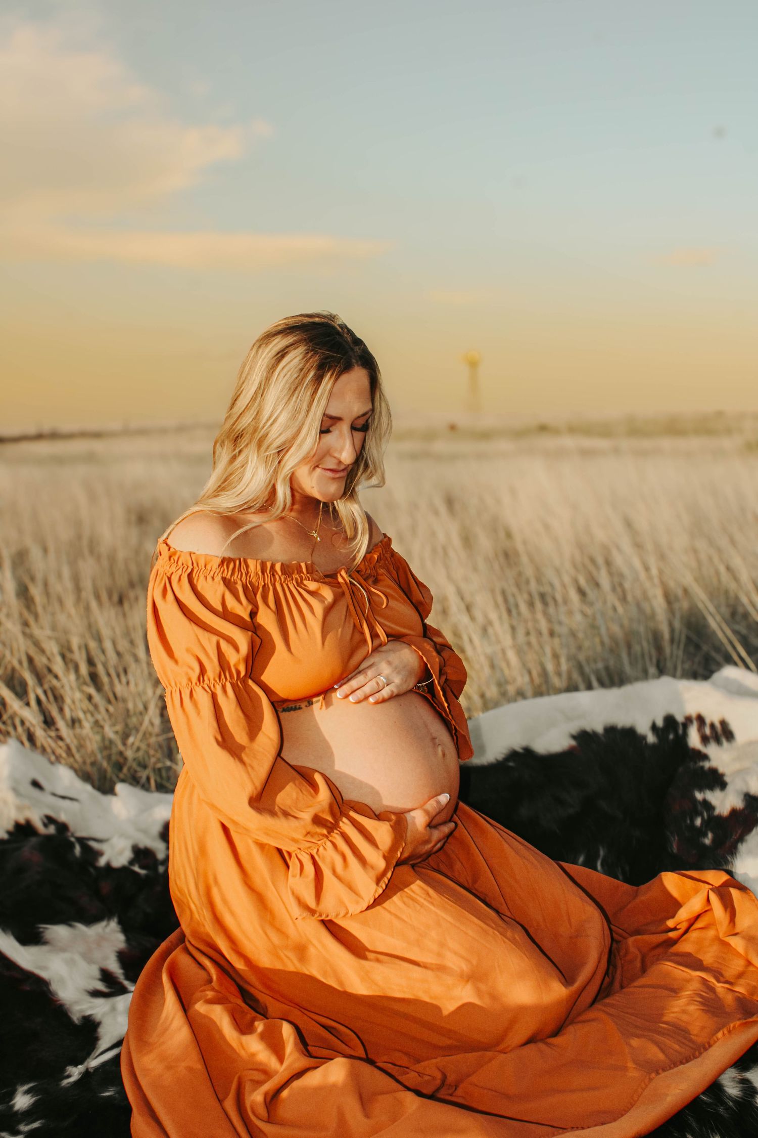 A woman in a flowing orange dress poses in a snowy field during sunset.