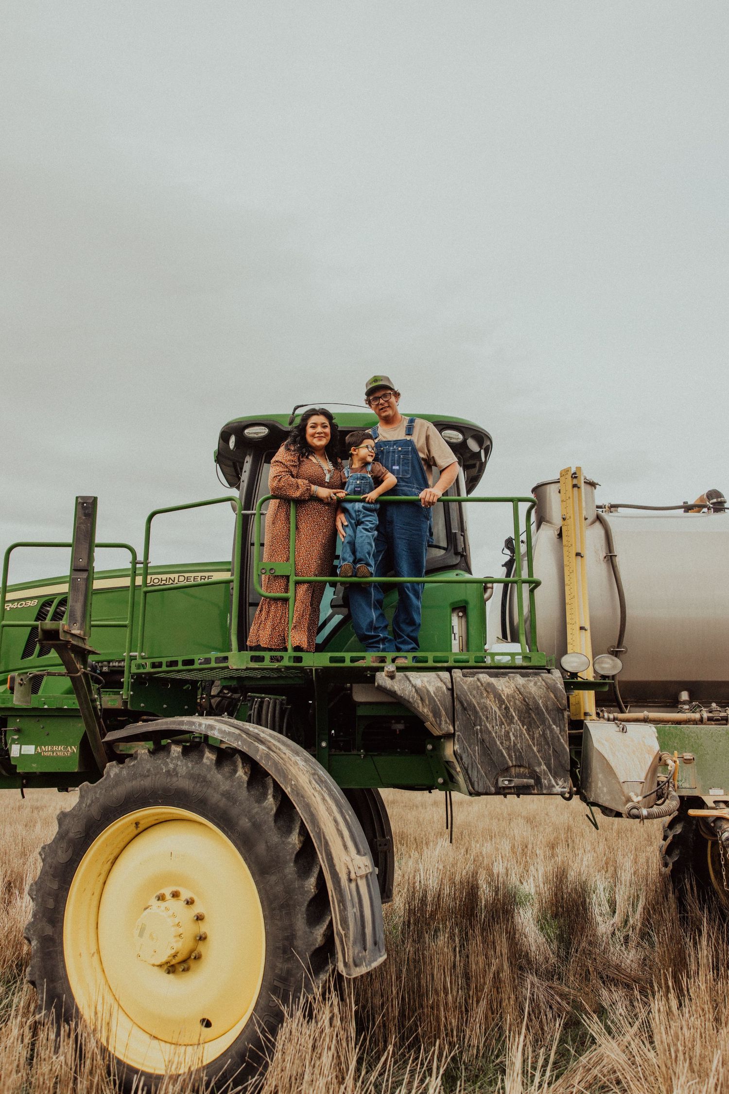 People ride in a large green John Deere tractor through a field of tall dry grass at sunset.