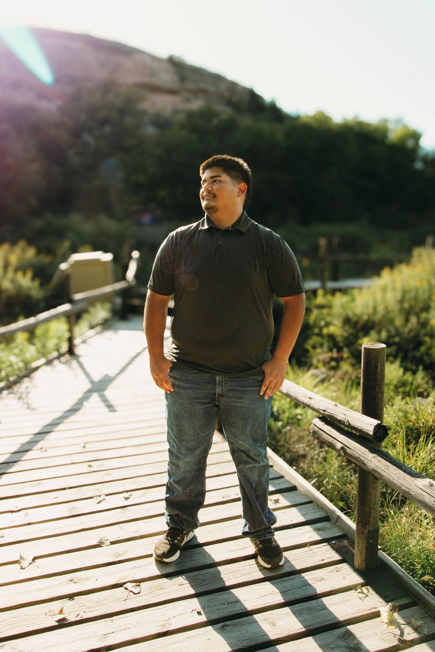 A person in a black polo shirt and jeans stands on a wooden boardwalk path during golden hour sunlight.