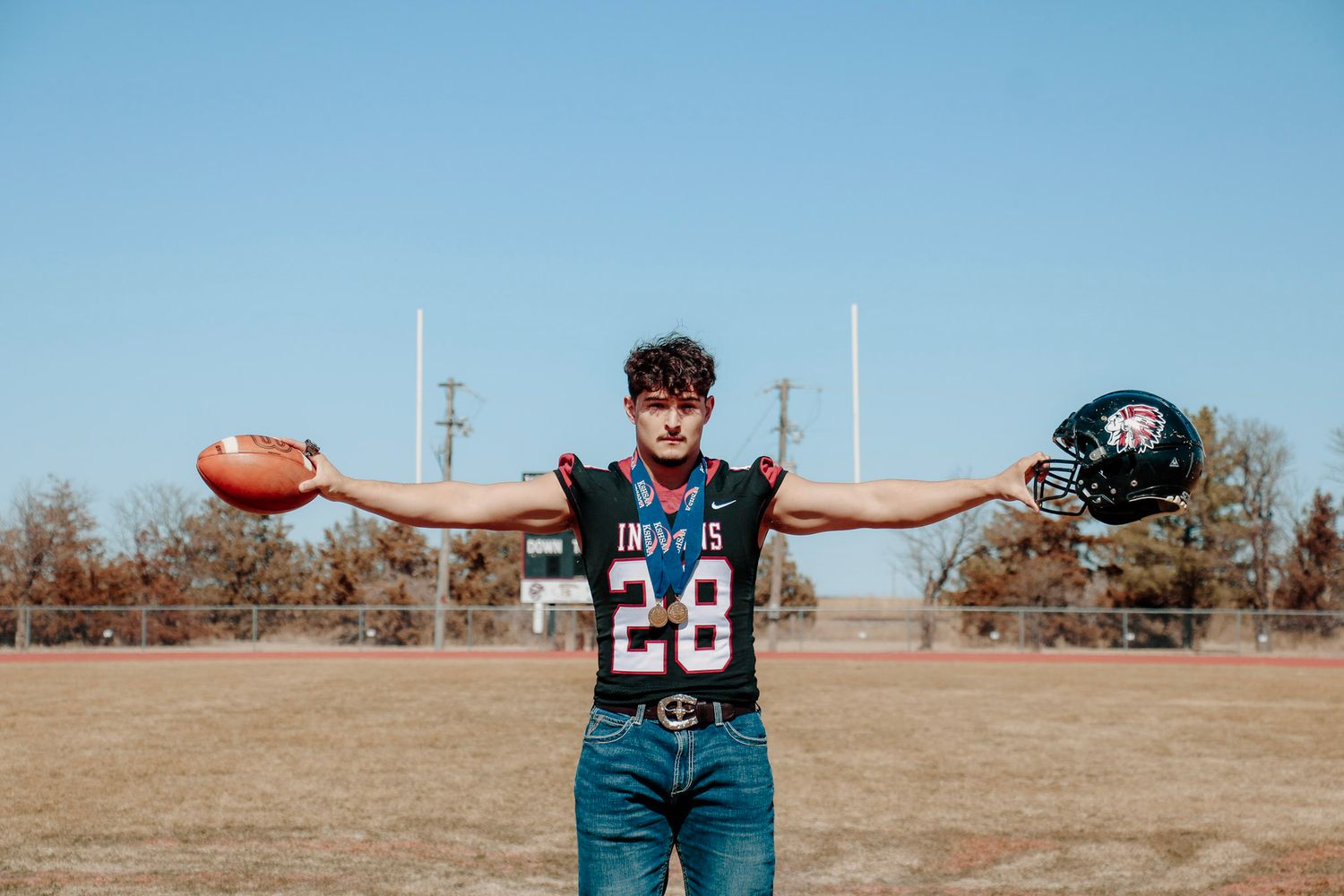 A person stands in an open field with arms outstretched, wearing a football jersey and jeans on a sunny day.