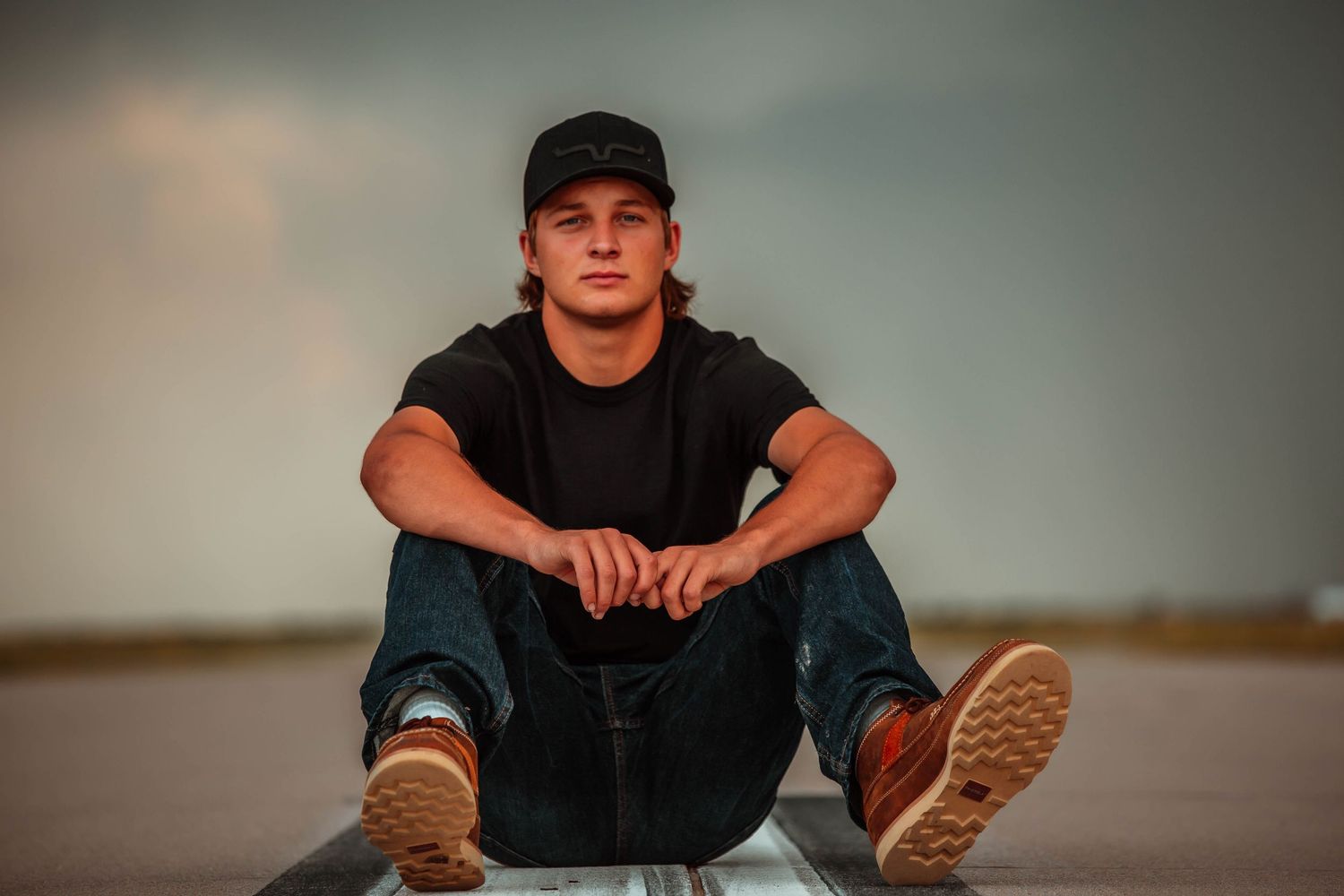 Person in black t-shirt and cap sits on wooden floor in artistic portrait series with moody lighting.