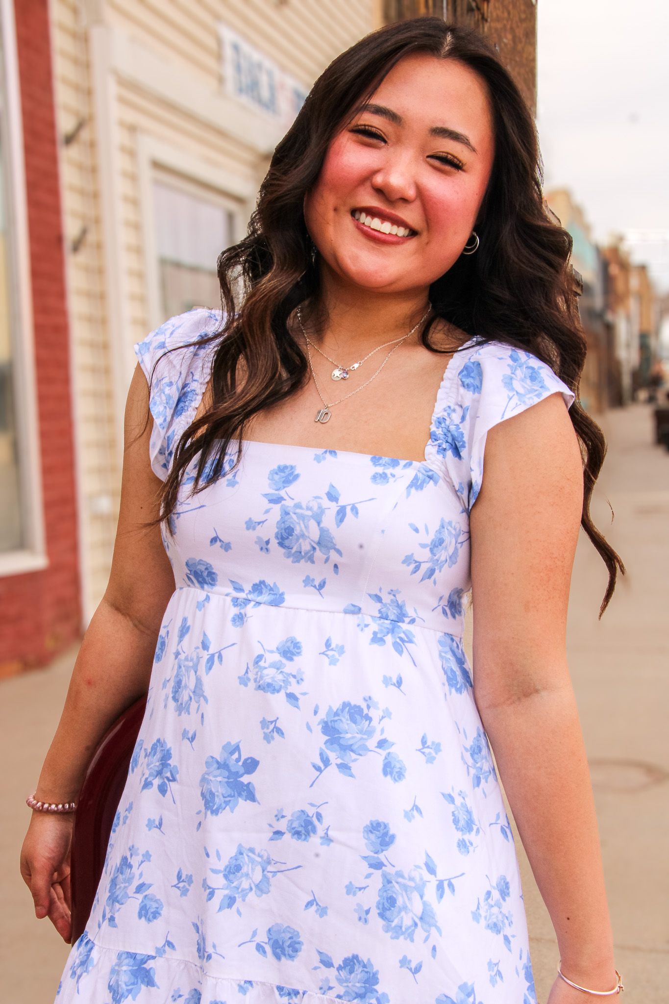 A floral dress in white and blue is shown in multiple angles on a sunny sidewalk against a brick wall.