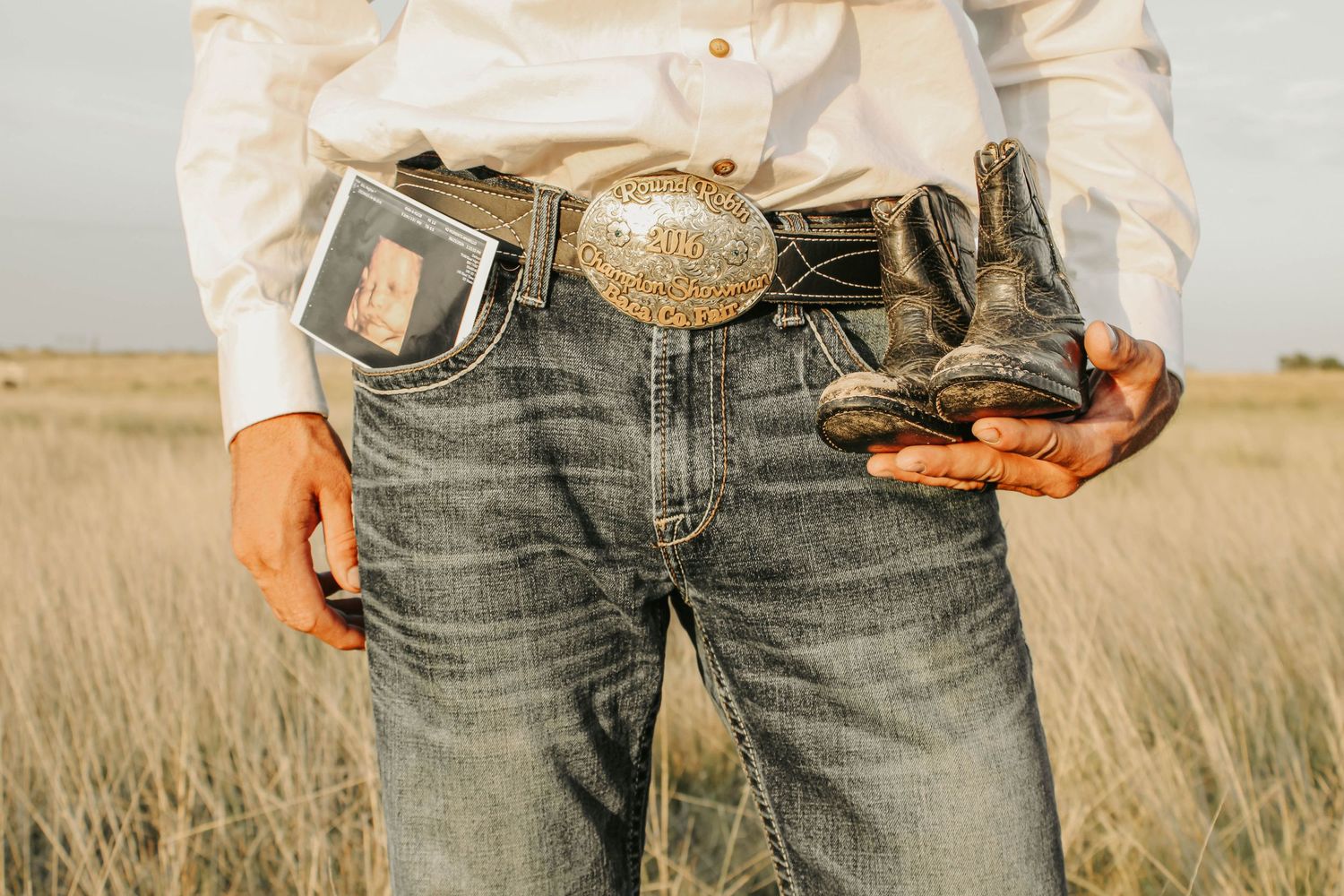 Close-up of western style jeans with an ornate belt buckle and boots against a prairie backdrop.