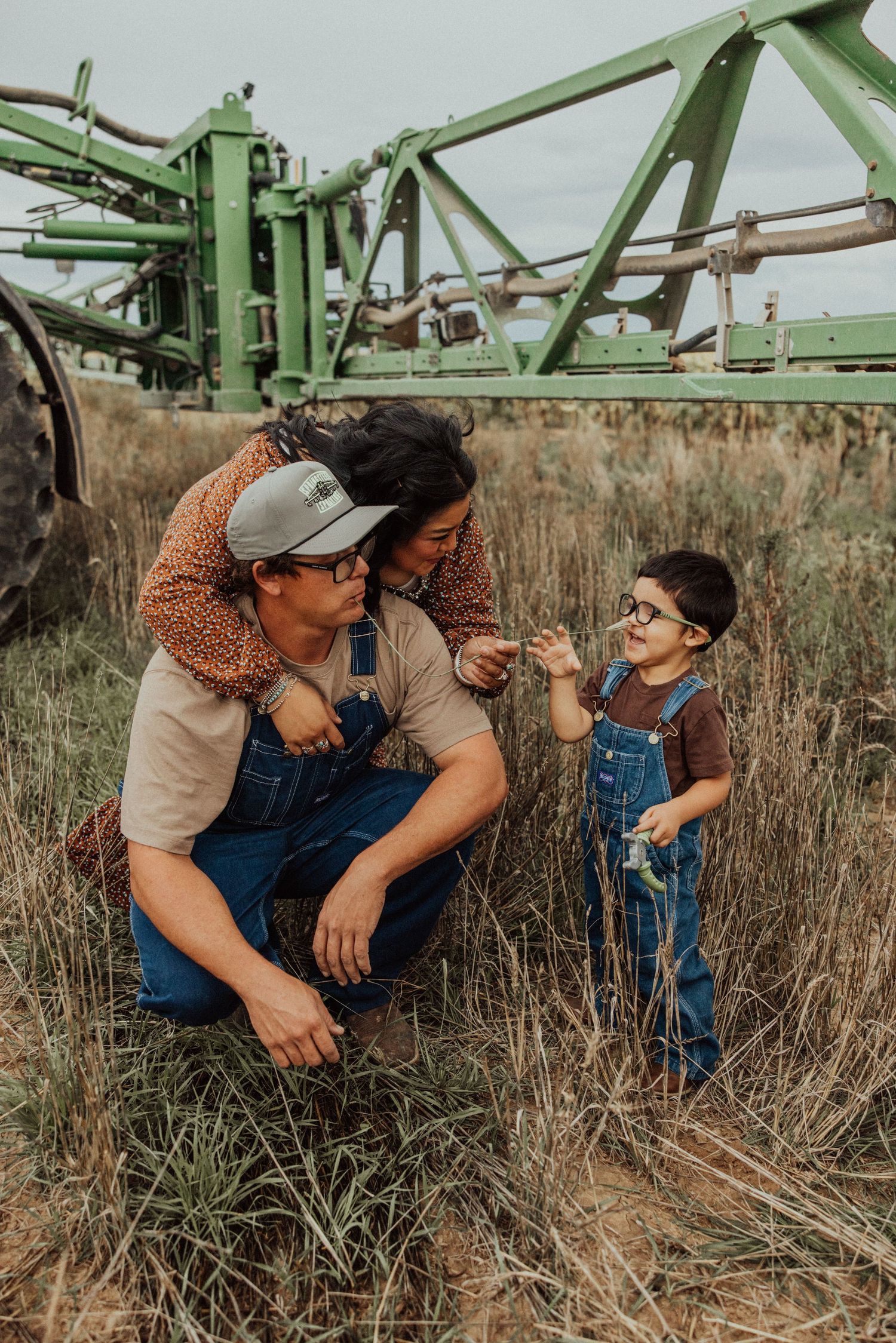 Two people in matching overalls and casual wear share a moment together in a field near farming equipment.