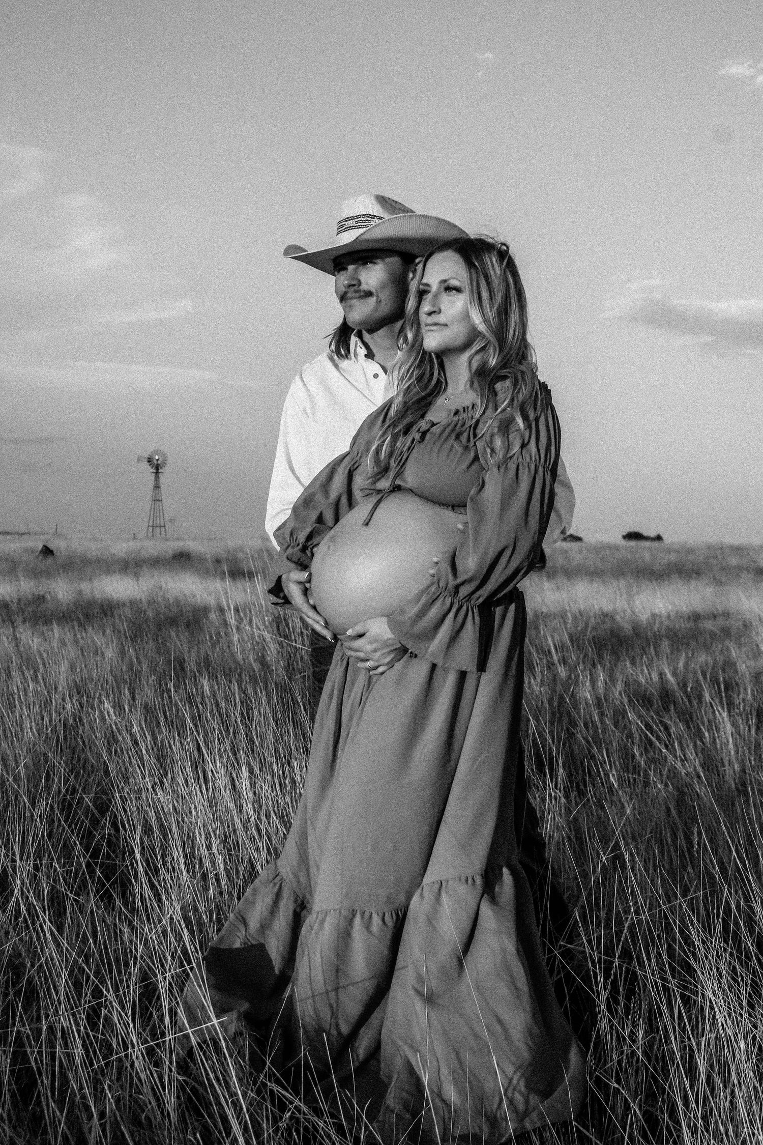 A couple in western attire poses together in a prairie field with a windmill in the background.