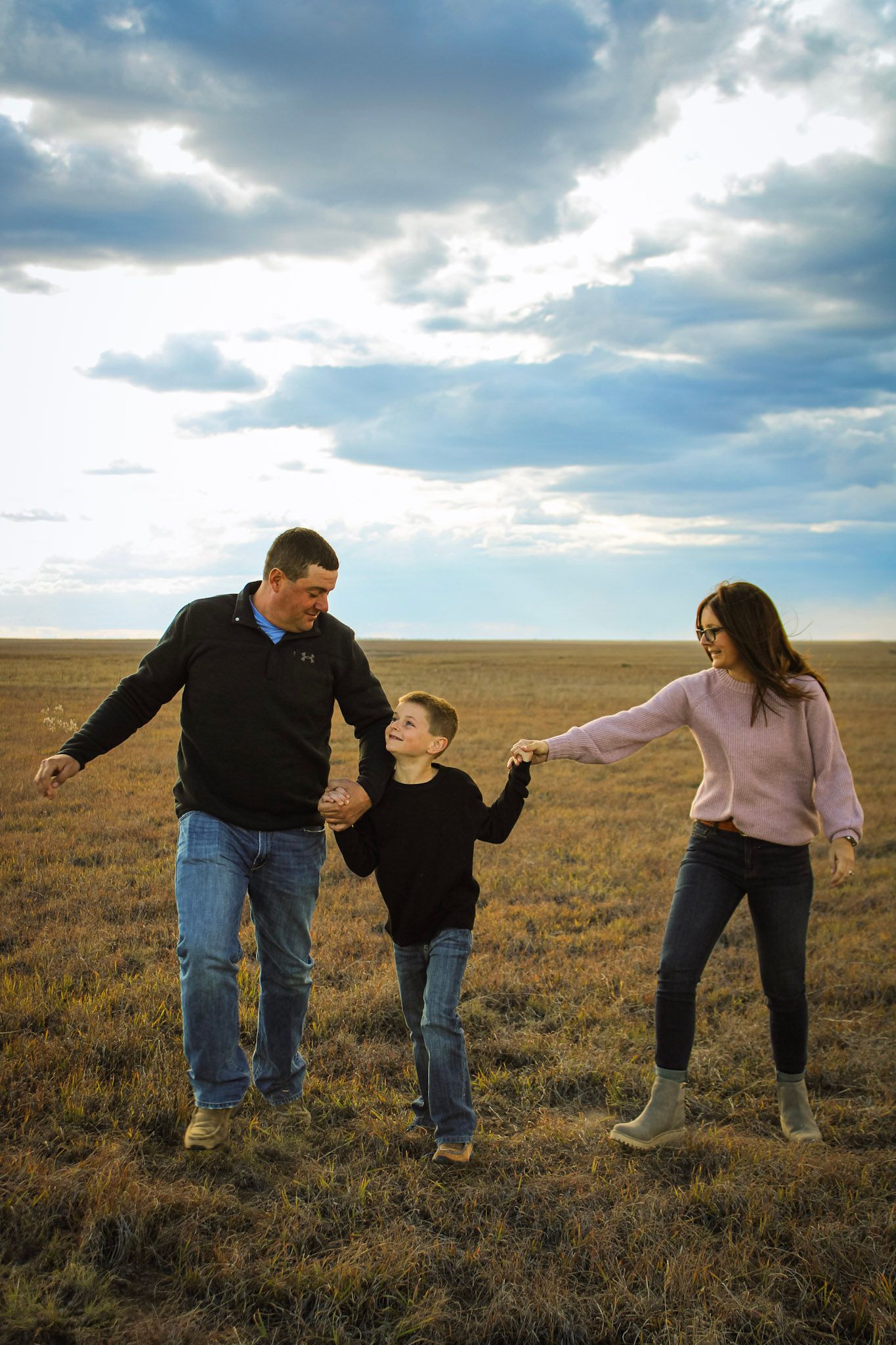 Family of three holding hands and playing together in an open field at sunset.
