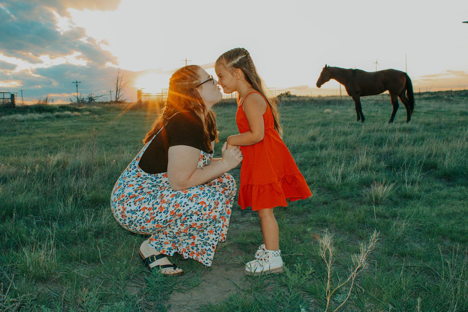 Two people in colorful dresses share a tender moment in a field at sunset with a horse in the background.