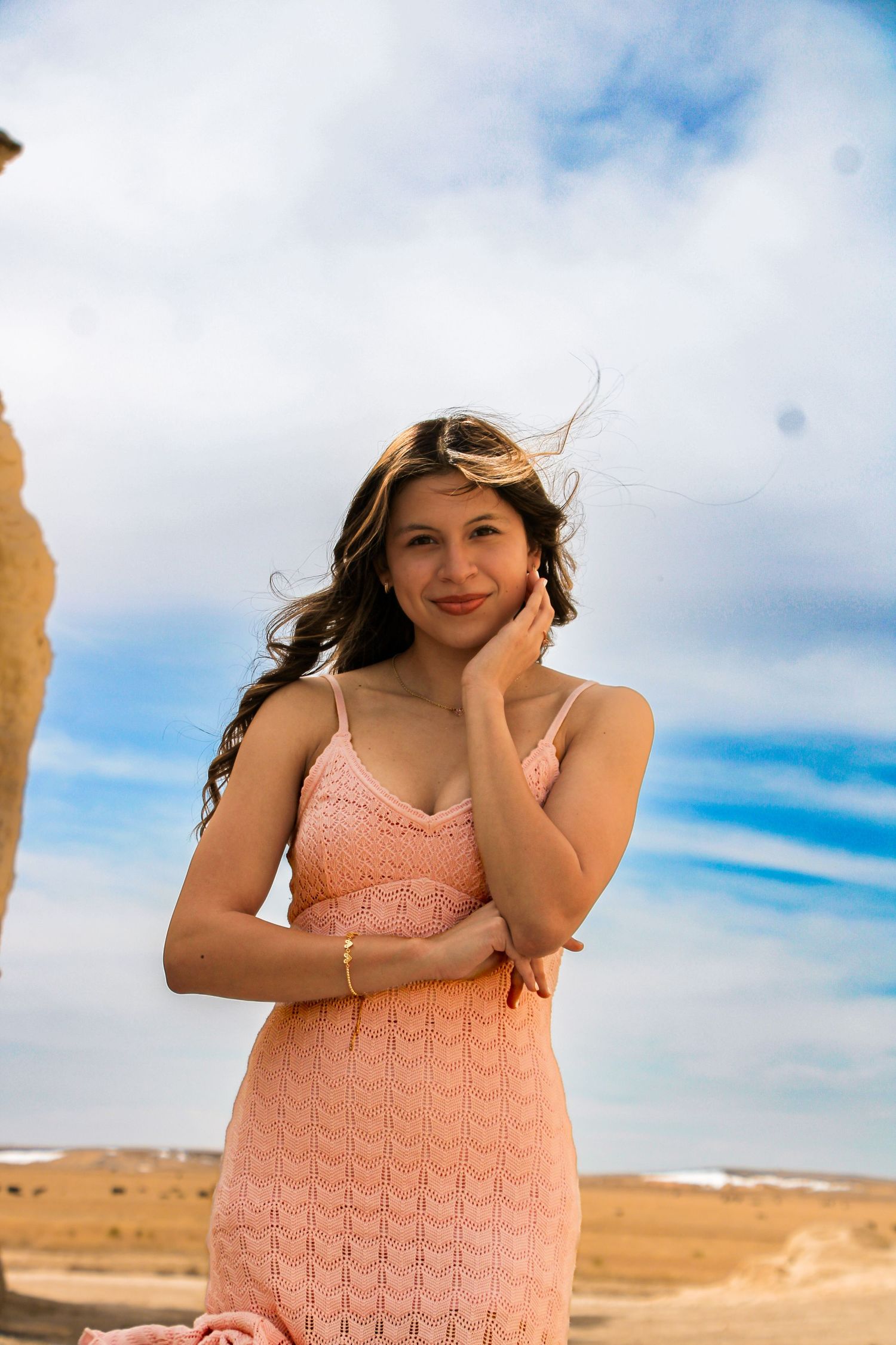 Person in peach-colored dress poses against desert landscape with blue sky and clouds.