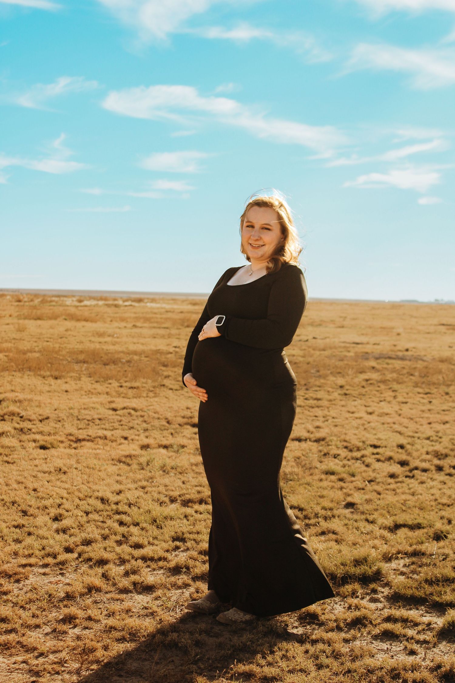 A pregnant figure in a flowing black dress poses confidently against a golden prairie landscape under bright blue skies.