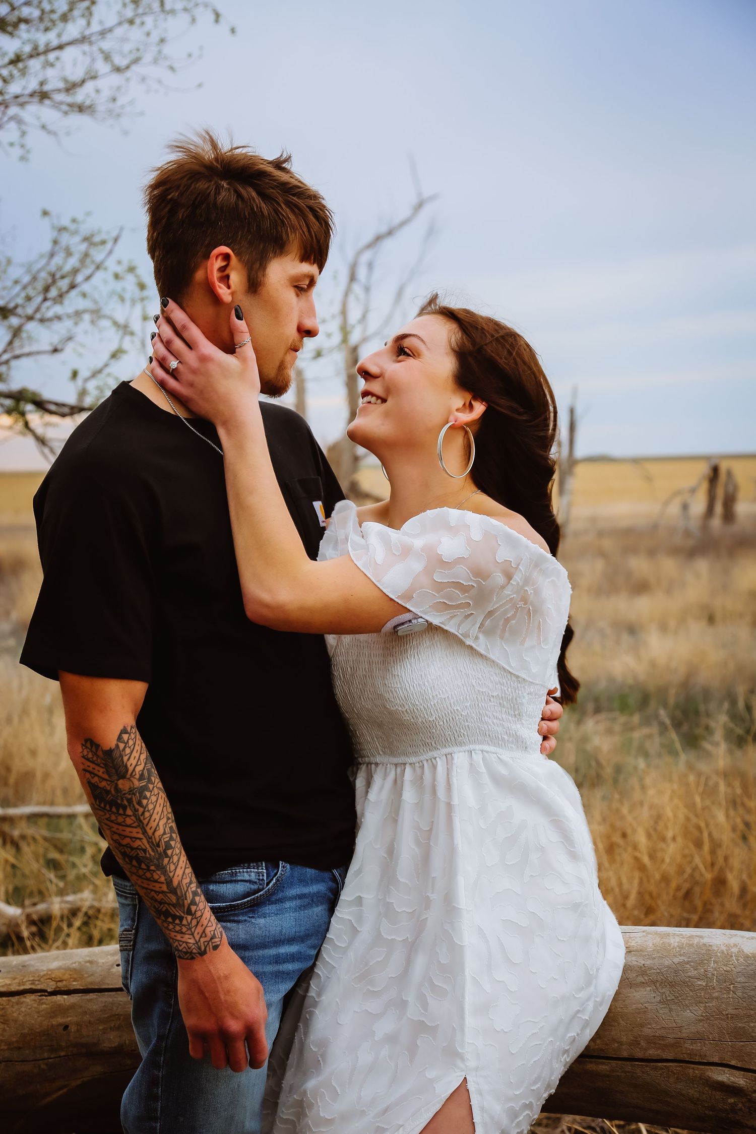 A romantic couple embraces on a fallen log in a rustic field with tall grass and cloudy skies.