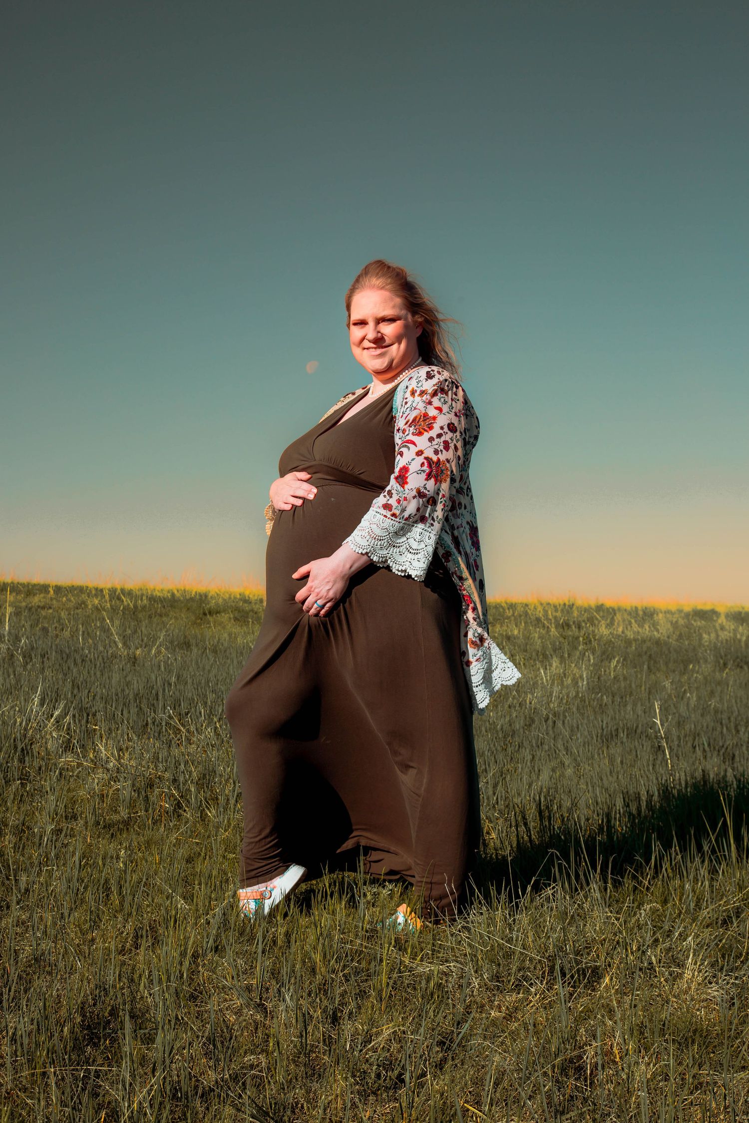 A pregnant figure in a brown maxi dress and floral cardigan poses in a grassy meadow at sunset.