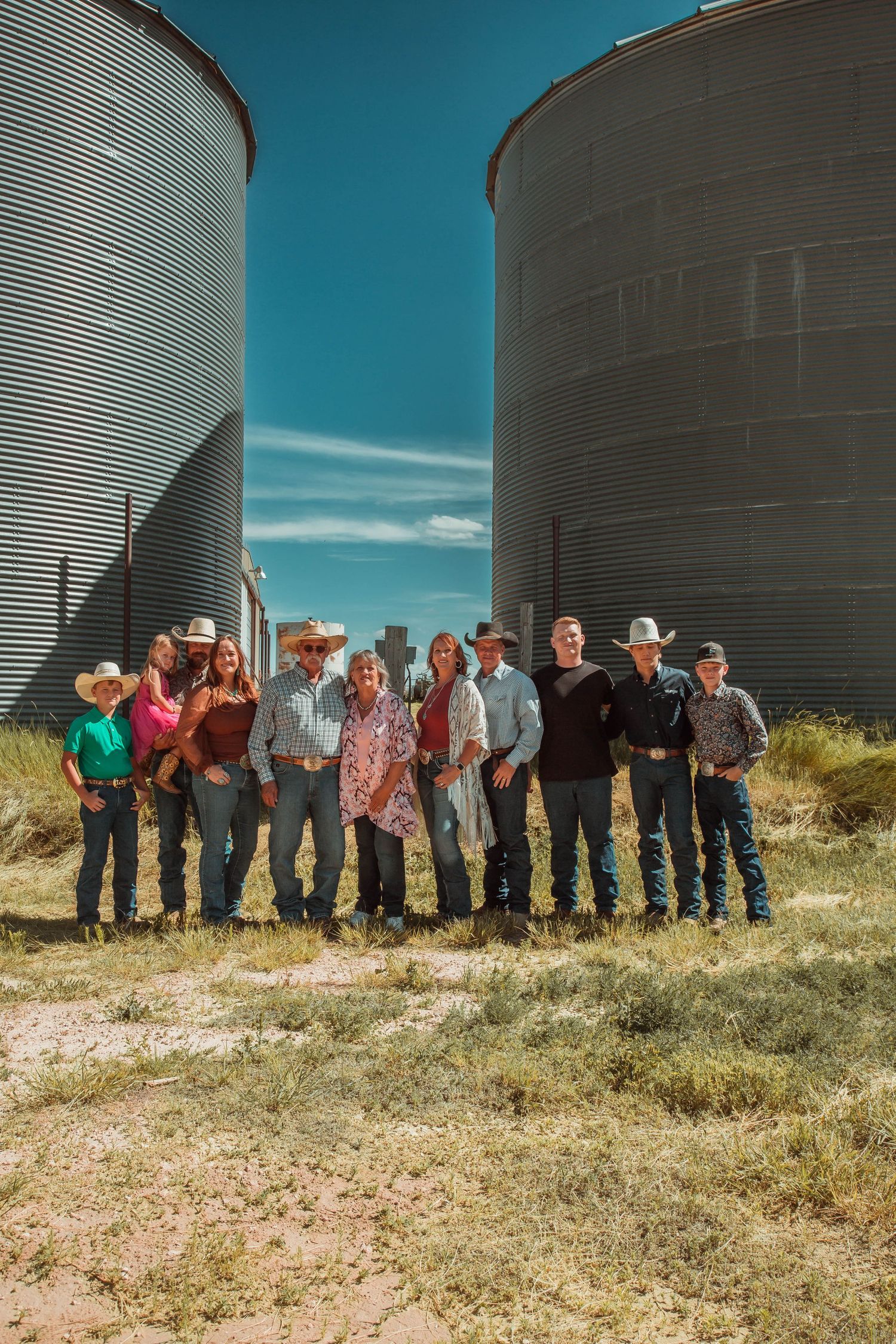 A group stands together on grassy ground in front of two large metal grain silos against a bright blue sky.