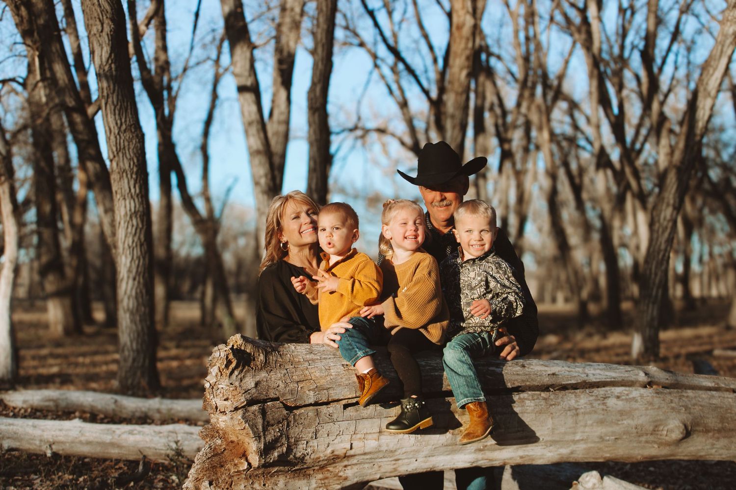 Children sitting on a fallen log among bare trees on a sunny day.