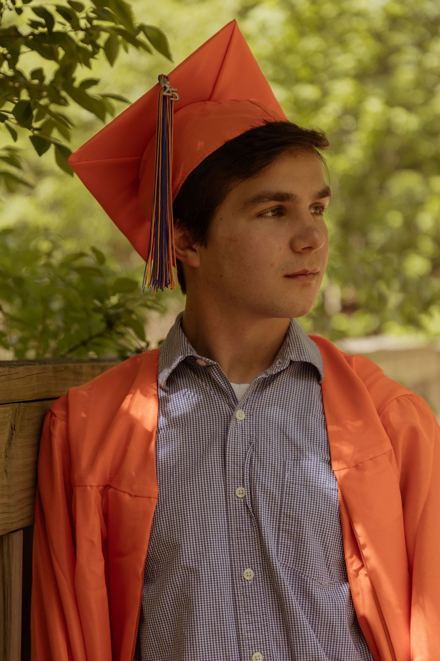 Graduate wearing orange cap and gown with striped dress shirt poses outdoors against blurred green foliage background.