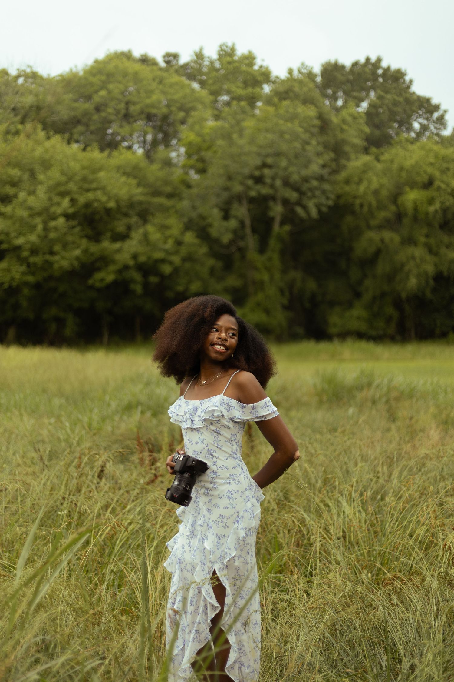 Smiling young Black woman with natural afro hair in floral off-shoulder dress holding camera in green meadow.