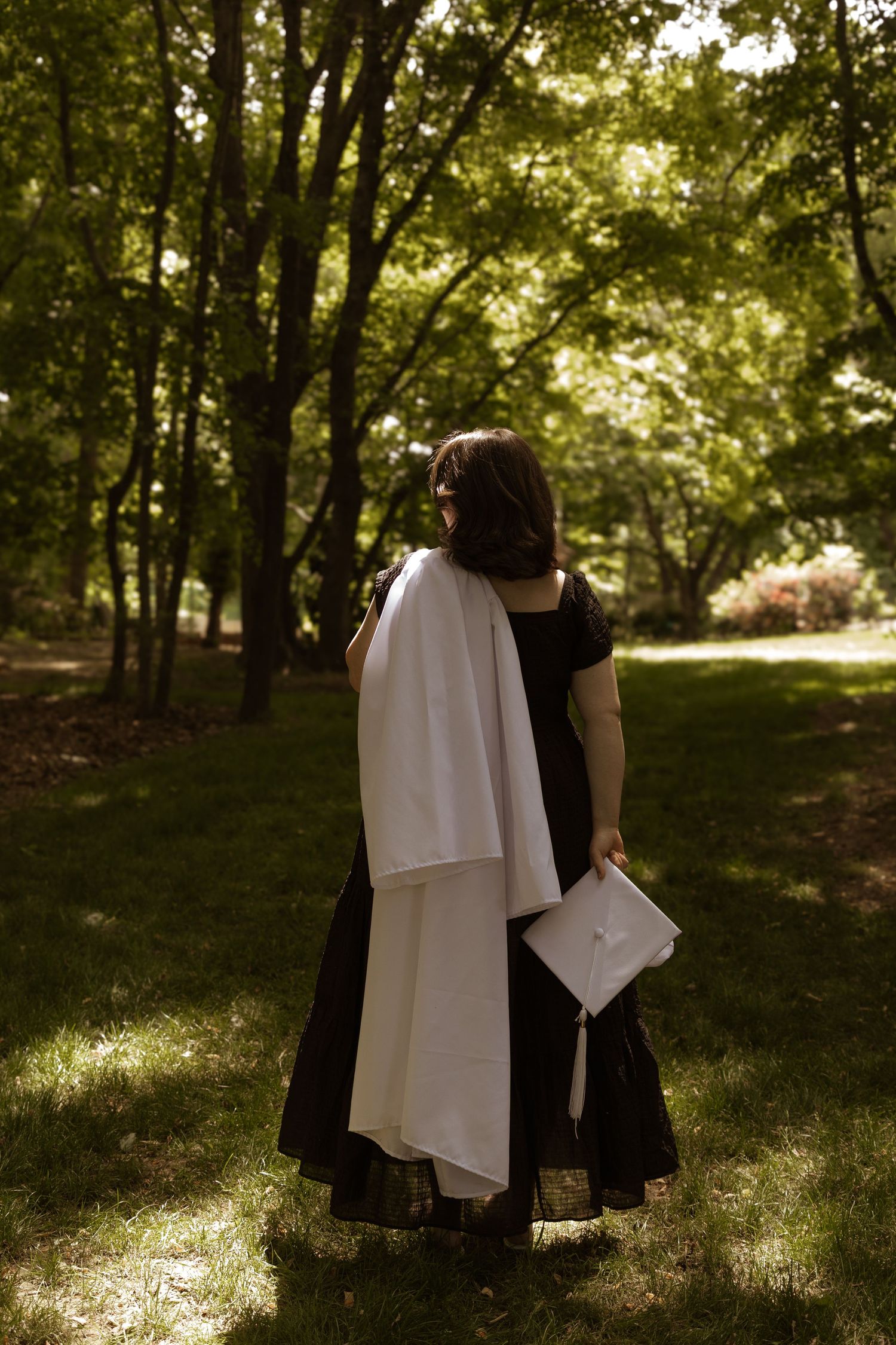 A figure in a black dress holds a white fabric and book while standing in a sunlit wooded area.