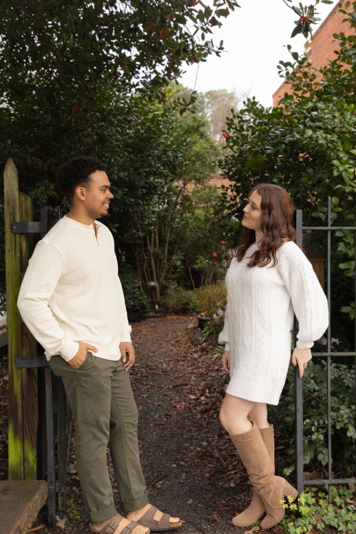 Two people in light sweaters chat while standing on a garden path surrounded by green foliage.