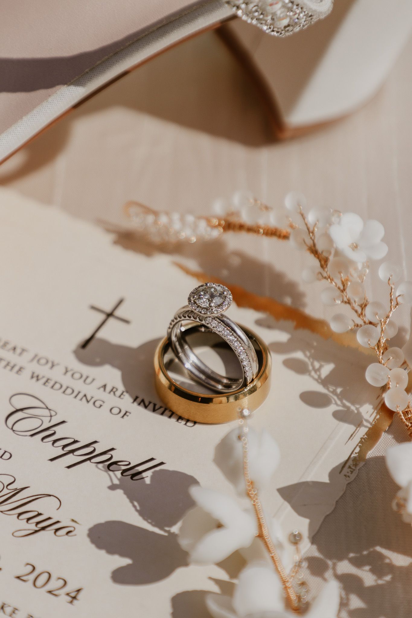 Wedding rings and invitation placed artistically with delicate white floral details and shadows.