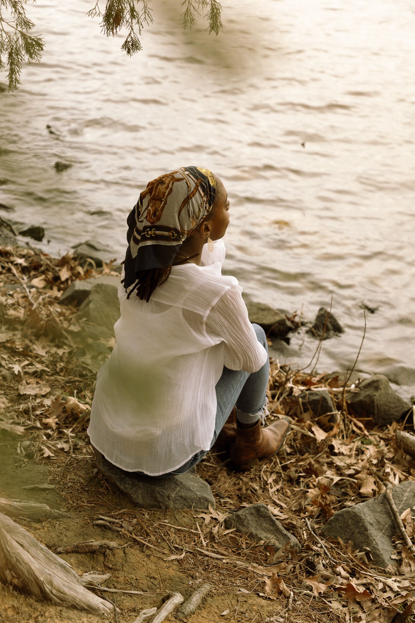 Figure sits on a rocky riverbank in a white shirt and headscarf, gazing contemplatively at the flowing water.