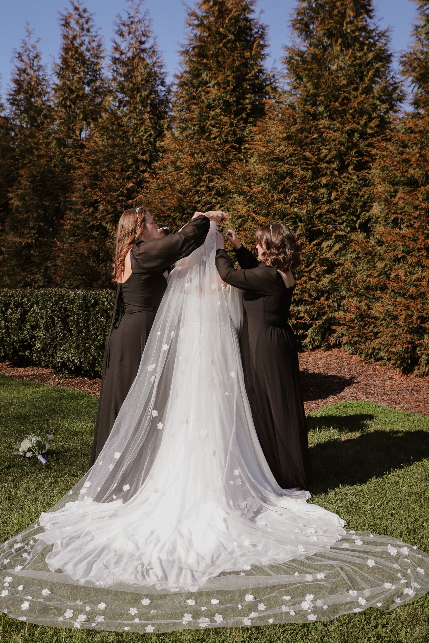 Bridesmaids in dark dresses adjust bride's long floral veil against evergreen trees.