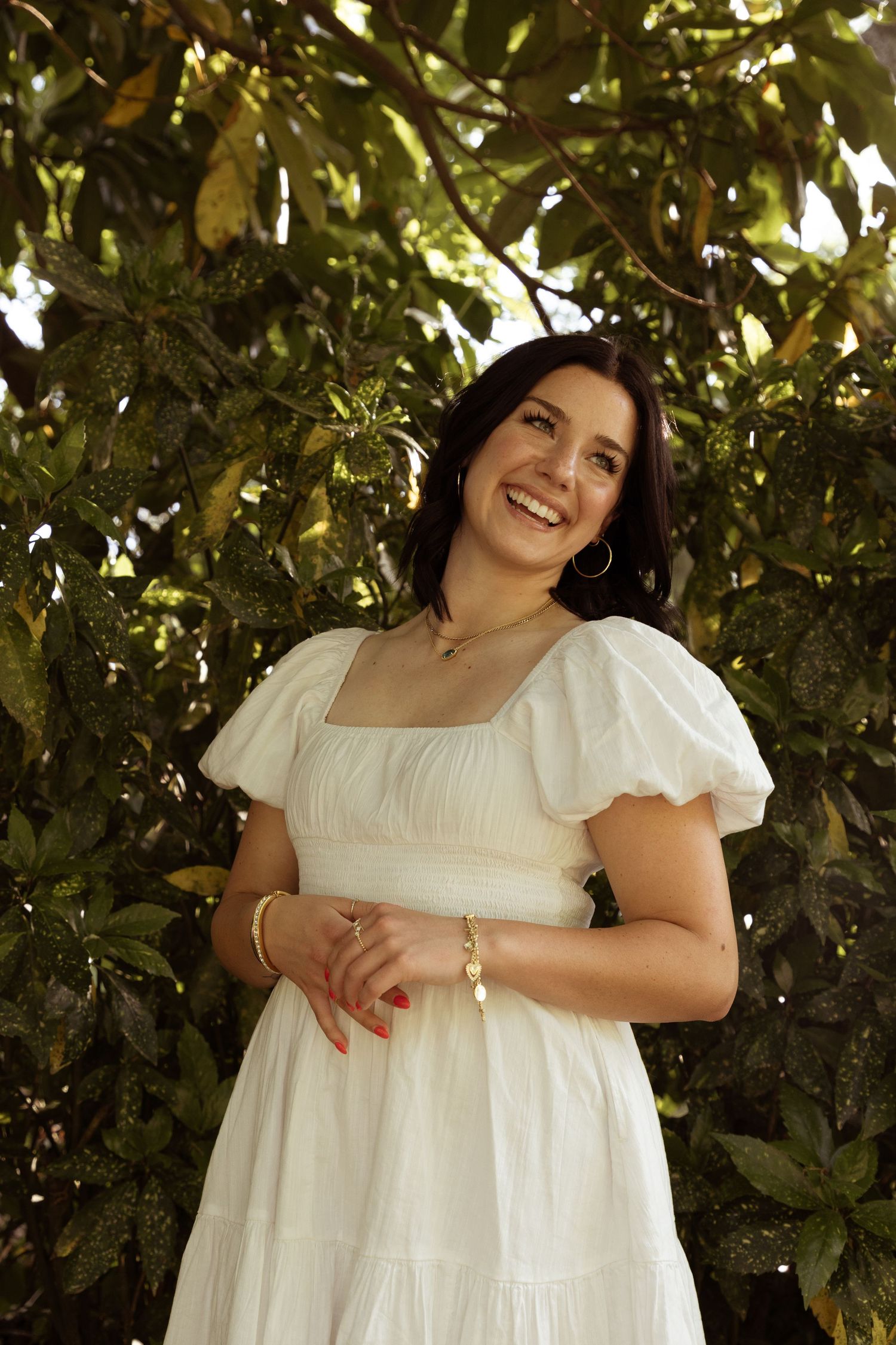 Graduate in white puff sleeve dress smiles brightly against leafy background.
