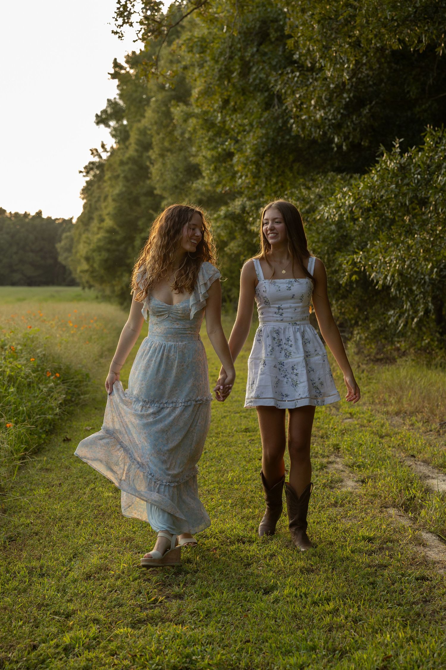 Two friends in flowing summer dresses walking along a grassy country path holding hands at sunset.