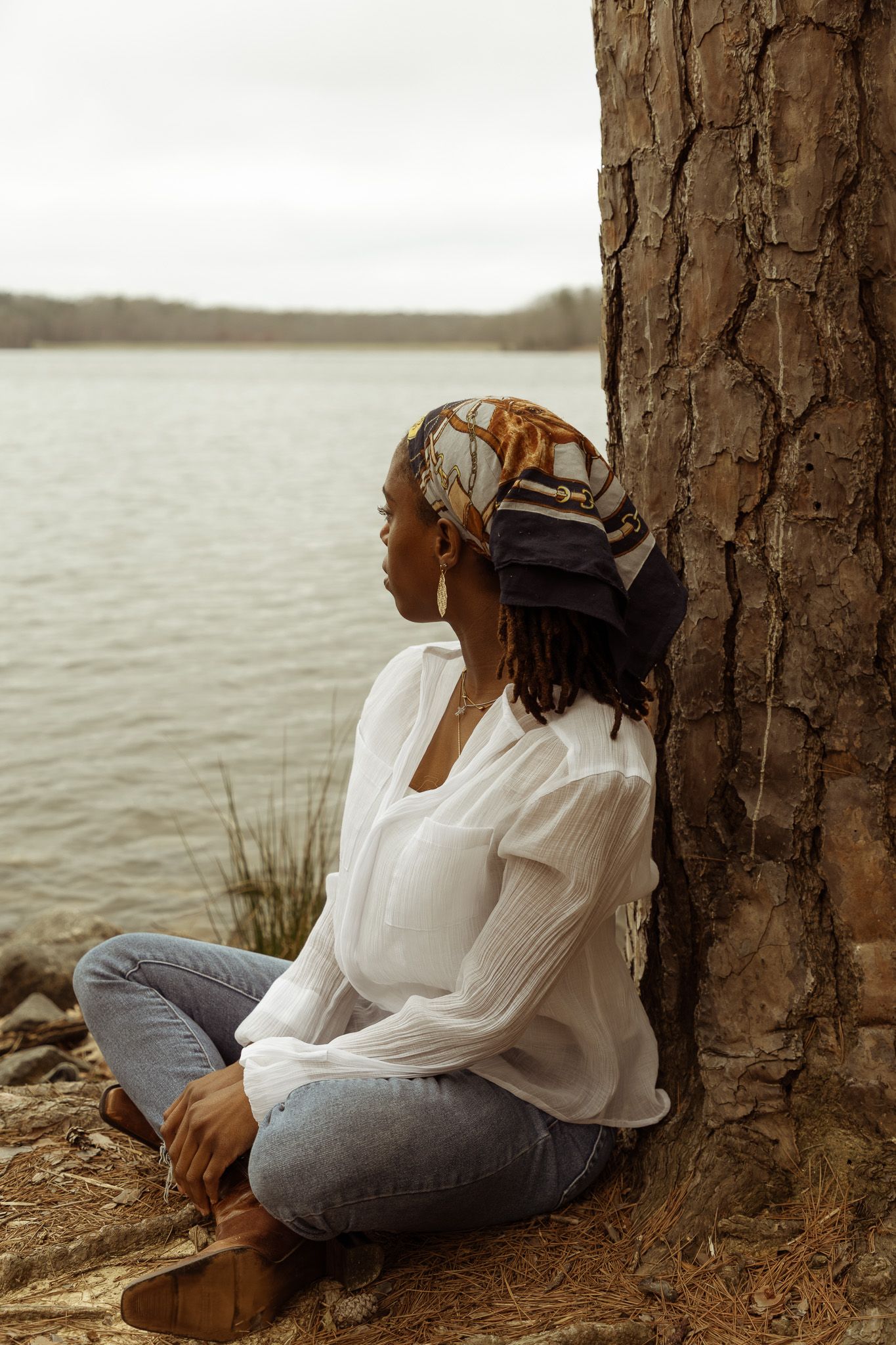 Person in white blouse and jeans sits peacefully against tree trunk by lake during golden hour.