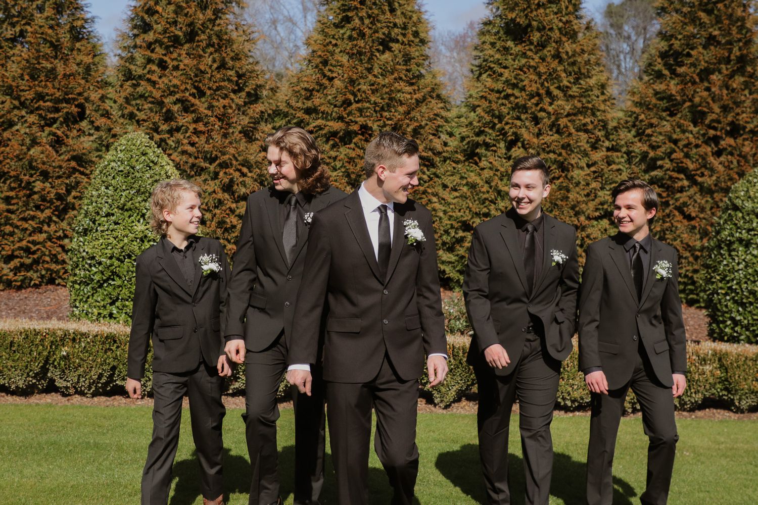 A group in formal black suits walks and laughs together along a green lawn with autumn trees in the background.