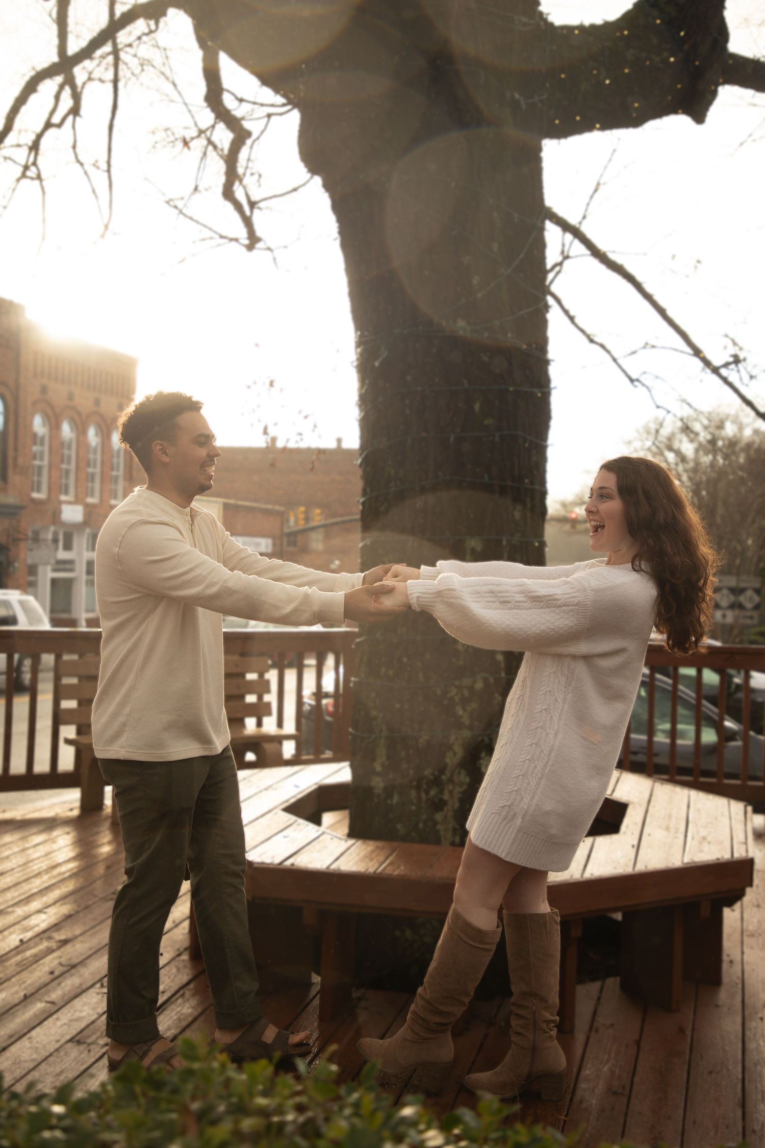 A couple holds hands while standing on a wooden deck near trees in autumn light while wearing cozy sweaters.