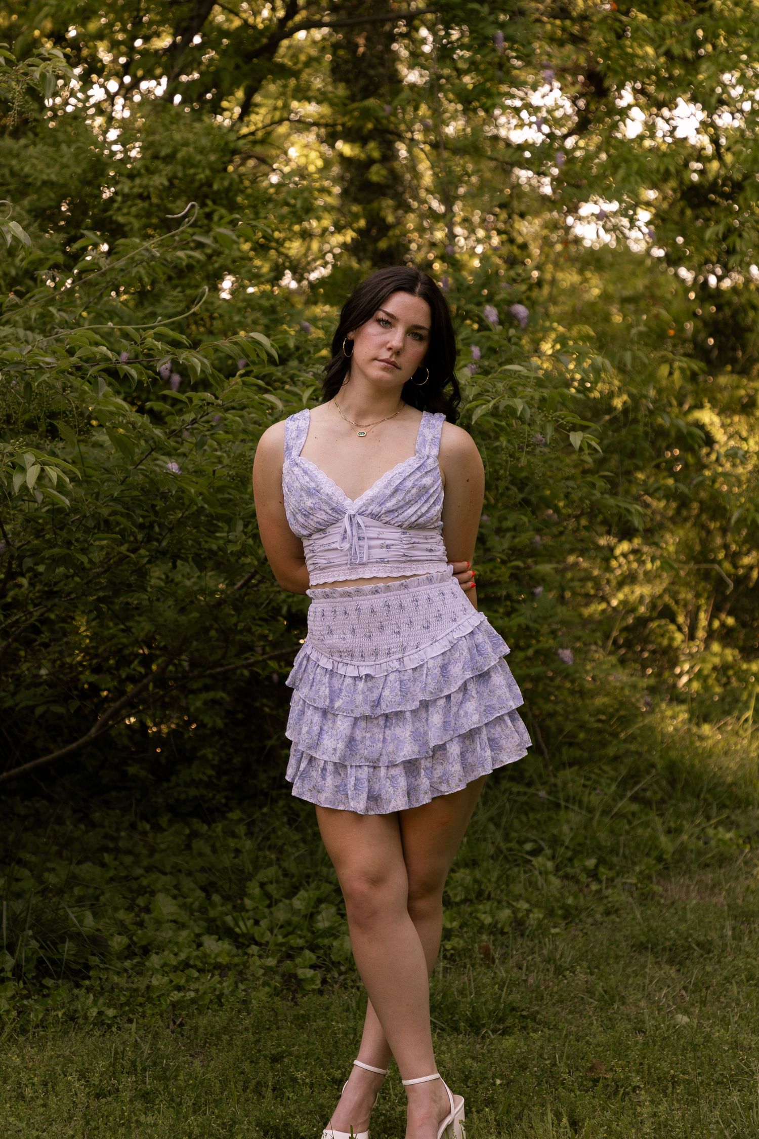 A person in a light lavender ruffled mini dress poses outdoors against a natural green backdrop at sunset.