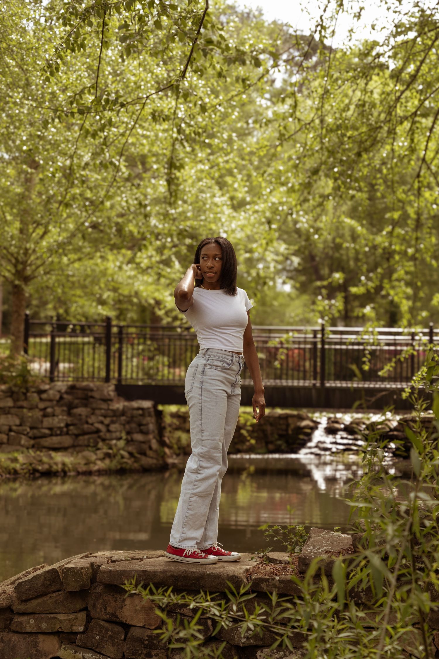 Person in white dress stands contemplatively by a tranquil pond with stone walls and footbridge in a serene park setting.