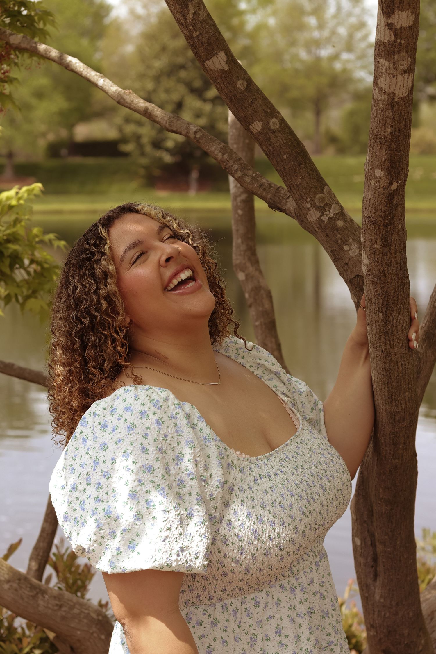 A joyful figure in a white floral dress leans against a tree trunk near a serene lake during golden hour.