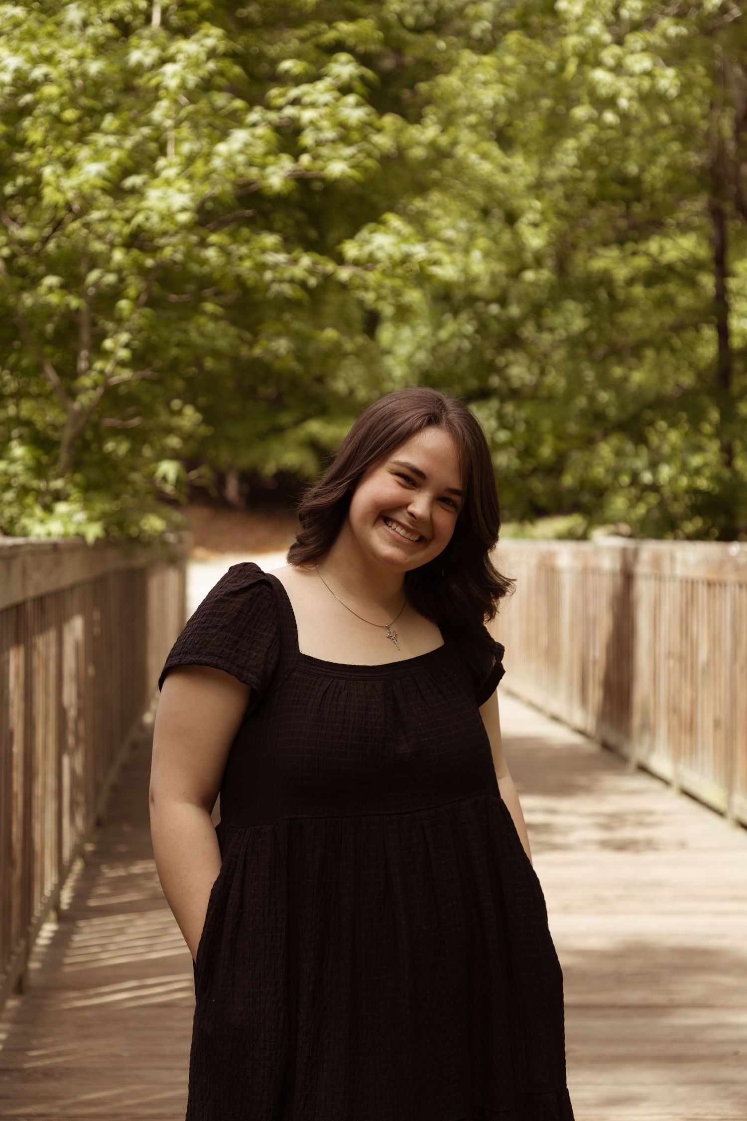 Dark-haired figure in a flowy black dress smiles warmly while standing on a wooden boardwalk surrounded by green foliage.