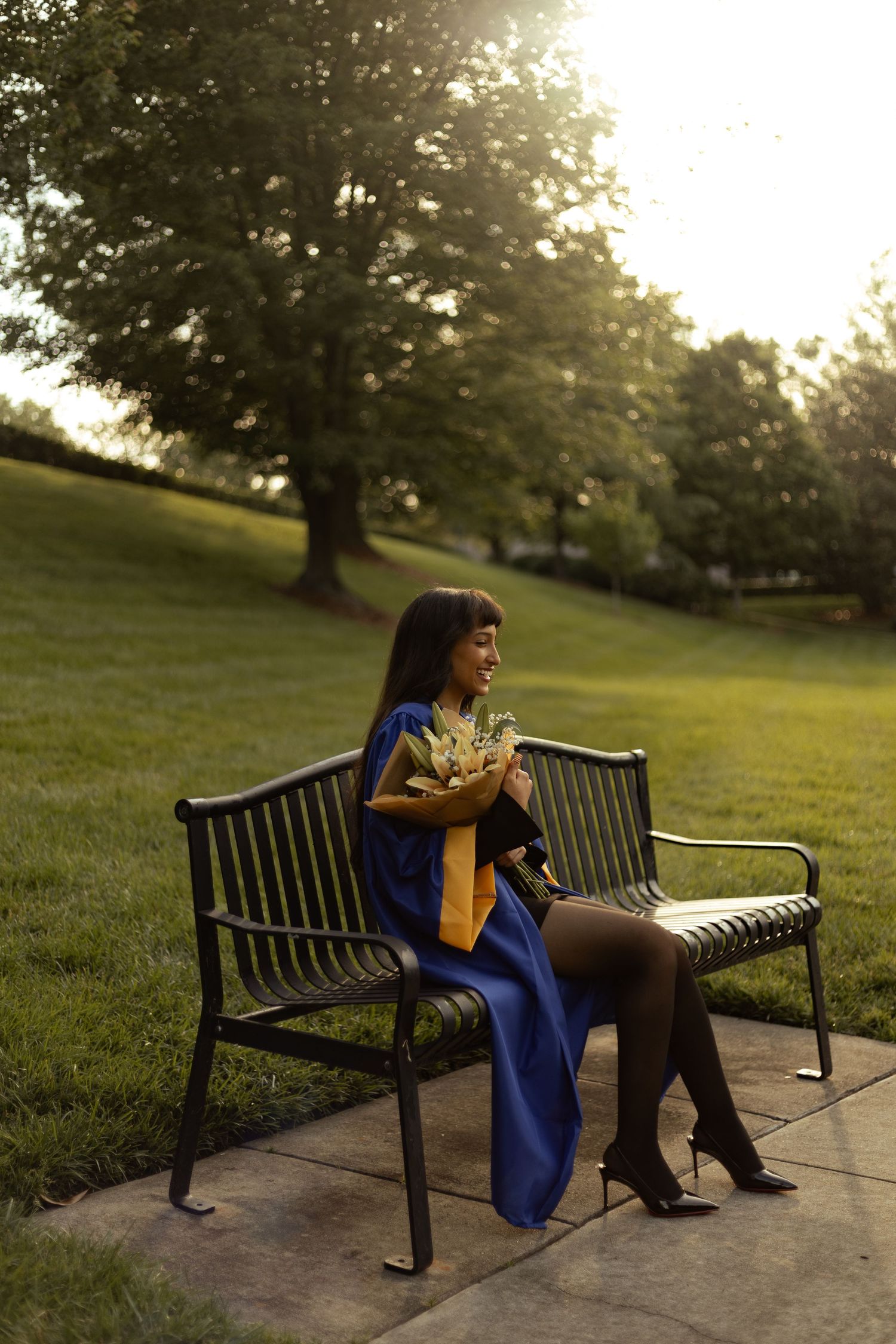 A young woman in blue graduation gown sits on a park bench holding flowers during golden hour sunset.