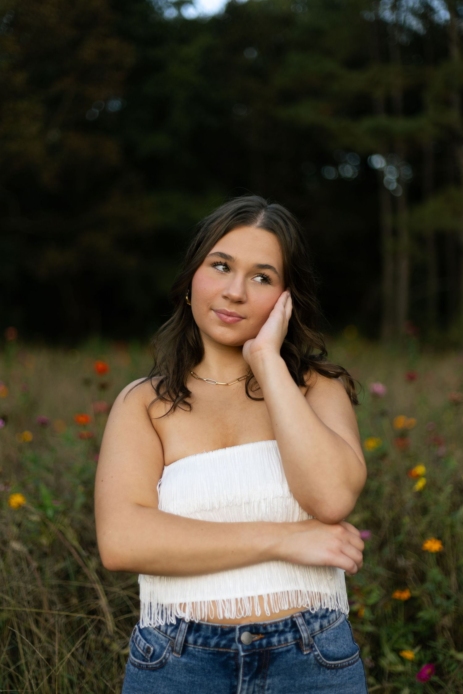 A person in a white fringe crop top and jeans poses thoughtfully in a field of wildflowers at sunset.