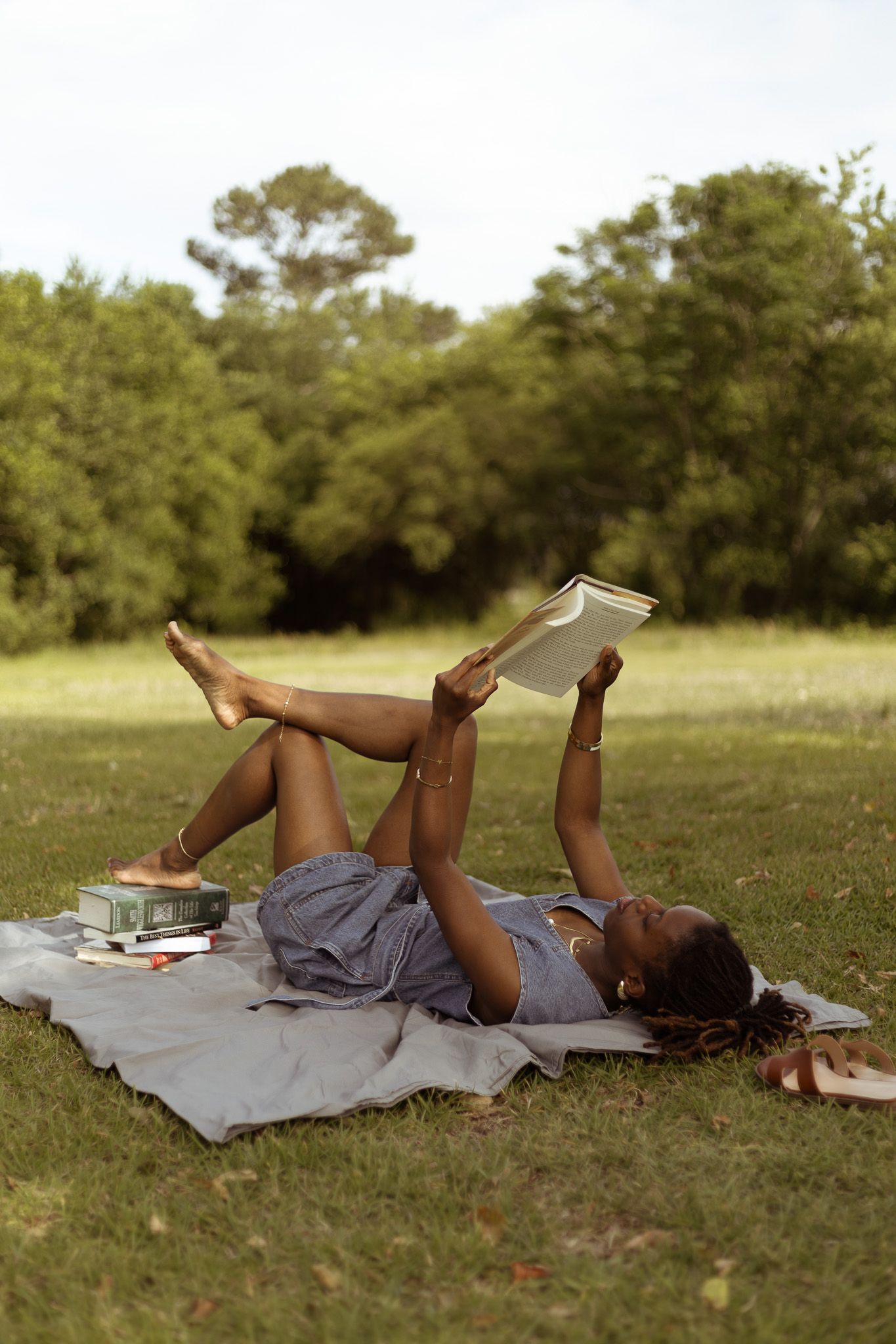 Someone relaxes on a blanket in a grassy field while reading a book on a sunny afternoon.