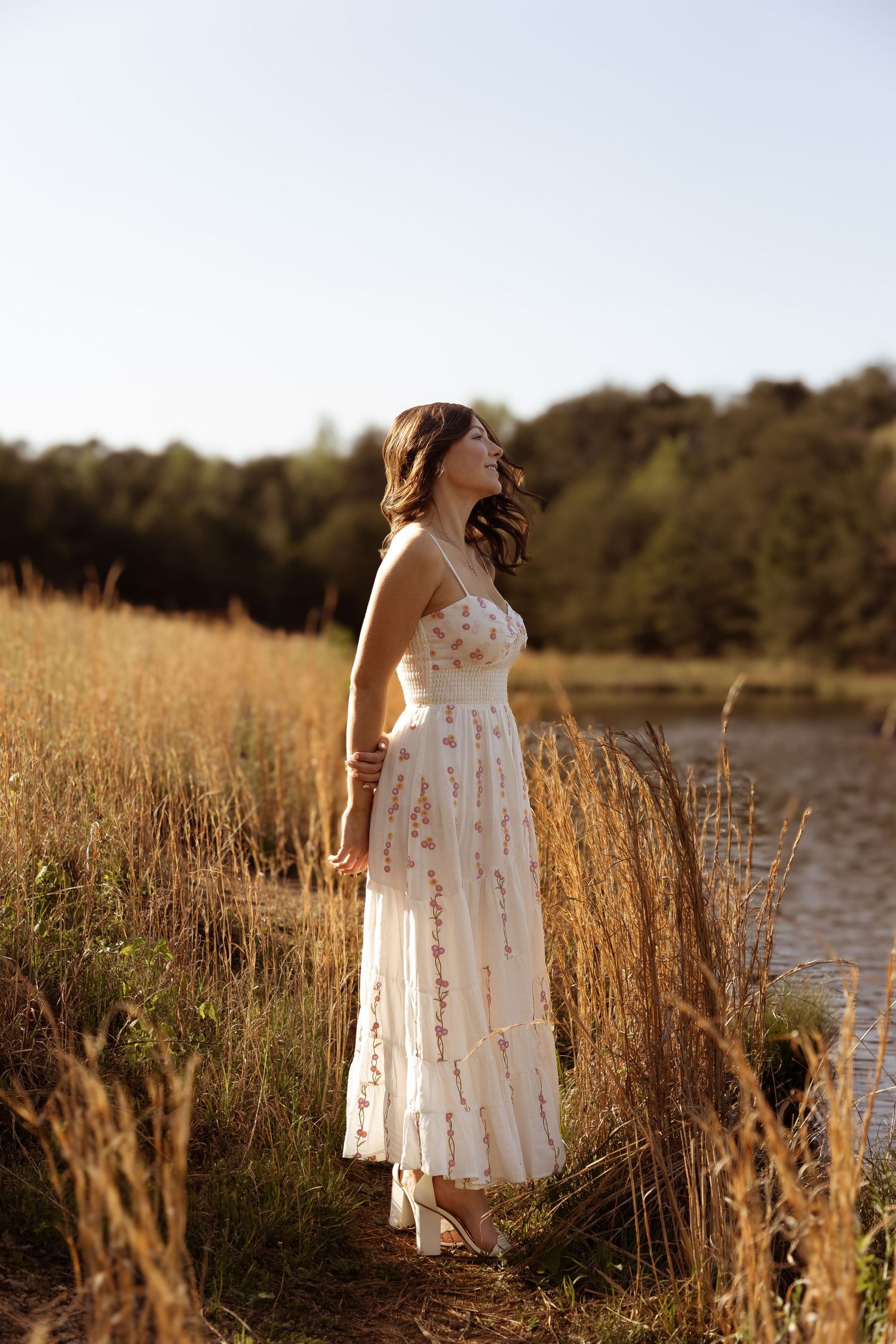 Graduate in white embellished dress poses among tall grass by waterfront during sunset.