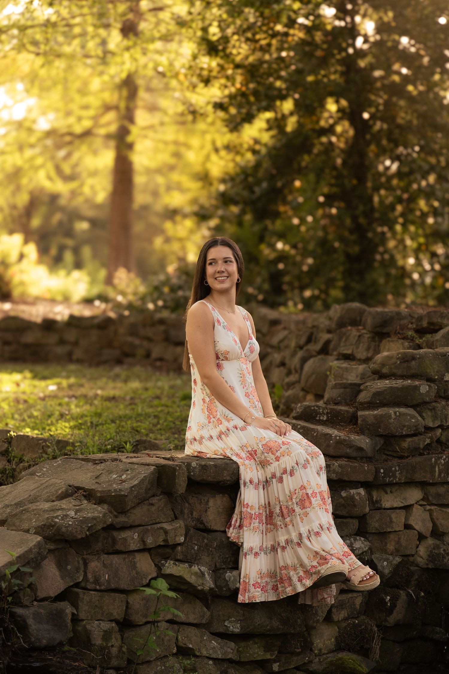 Woman in floral maxi dress sits on stone wall during golden sunset in garden.