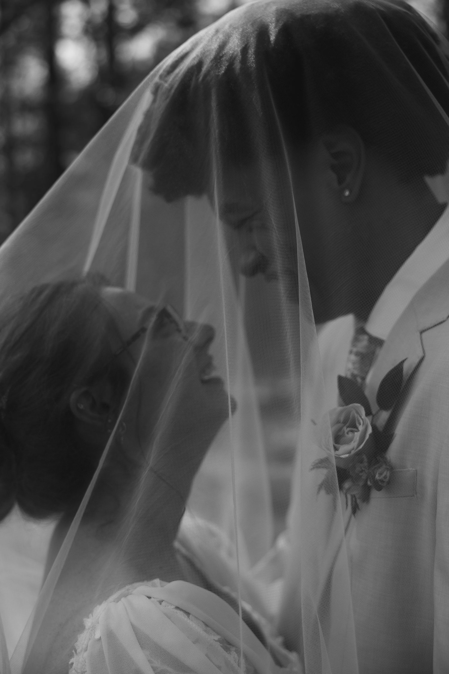 Black and white wedding photo of bride and groom sharing intimate moment under bridal veil outdoors.