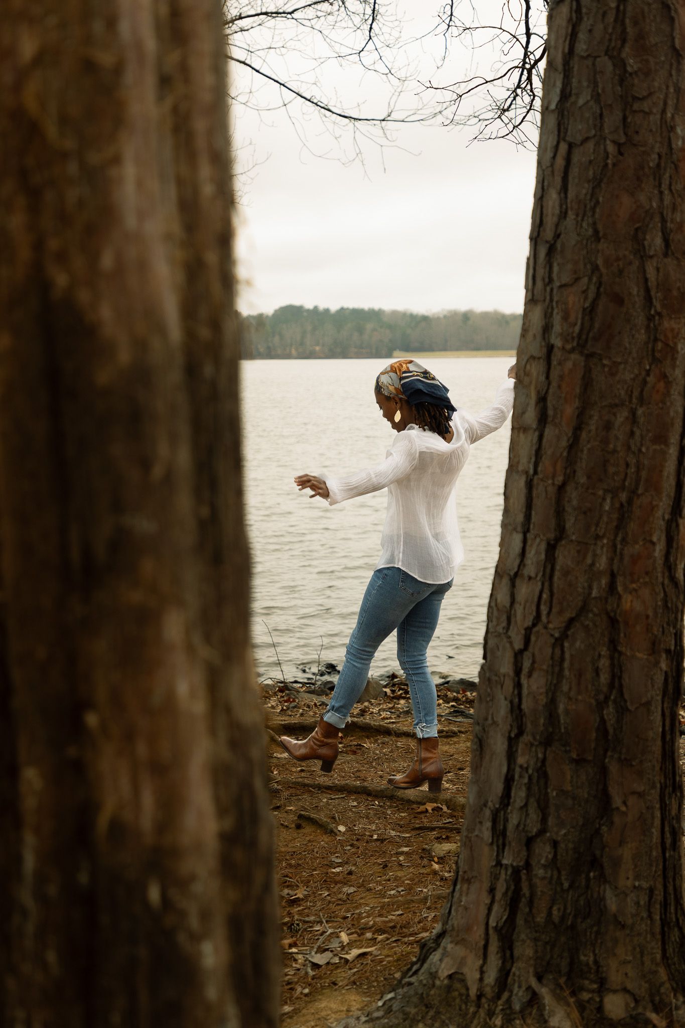 Person in white top and jeans dances playfully between trees along a scenic lakeside on an autumn day.