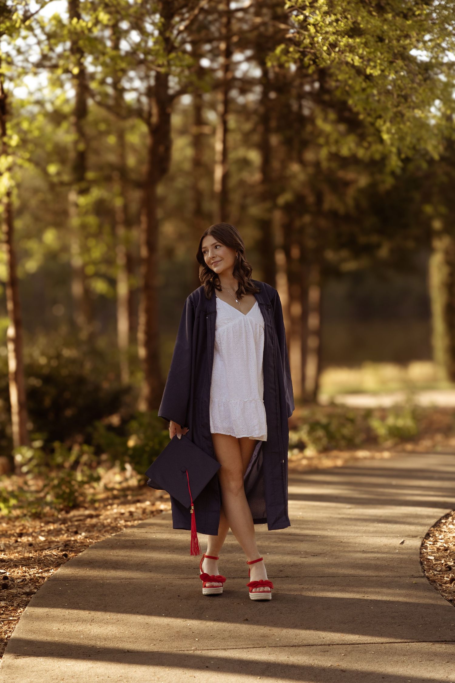 Person in a white dress and navy cardigan walks along a sunlit forest path wearing red sandals on a warm day.