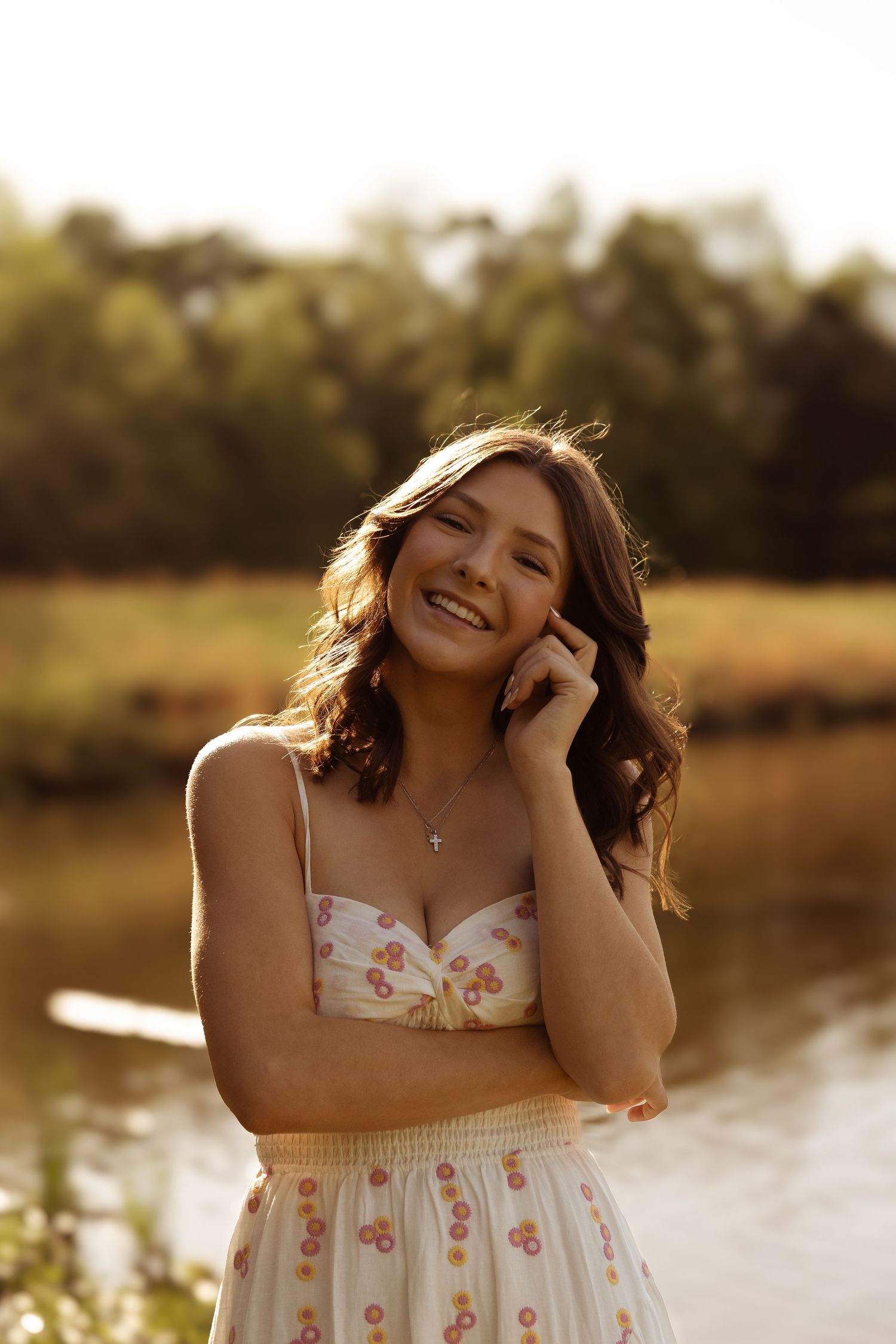 A young woman in a floral dress smiles warmly during golden hour near a pond, with sunlight creating a glowing effect.
