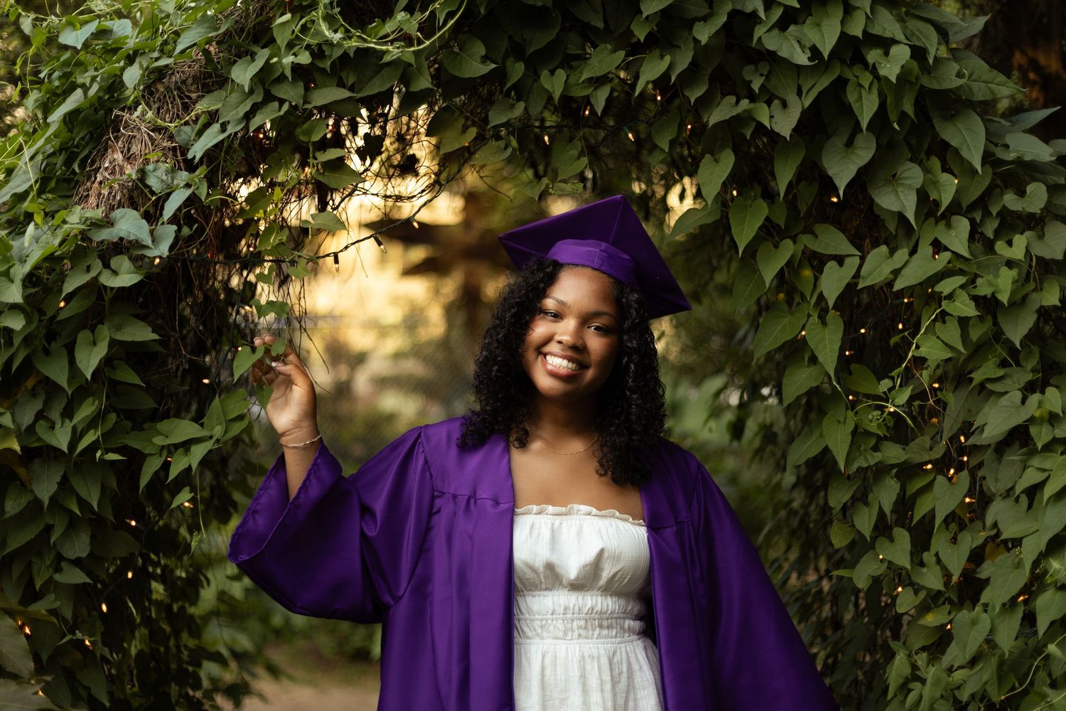 Smiling graduate in purple cap and gown posing outdoors among lush green ivy with string lights.